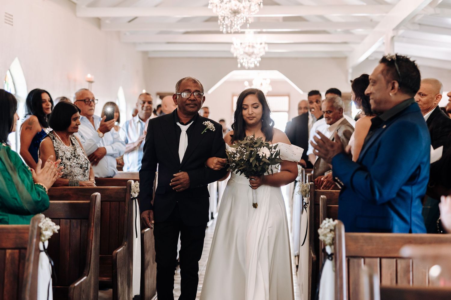 Guests celebrate and clap during a wedding ceremony in a bright chapel with wooden pews.
