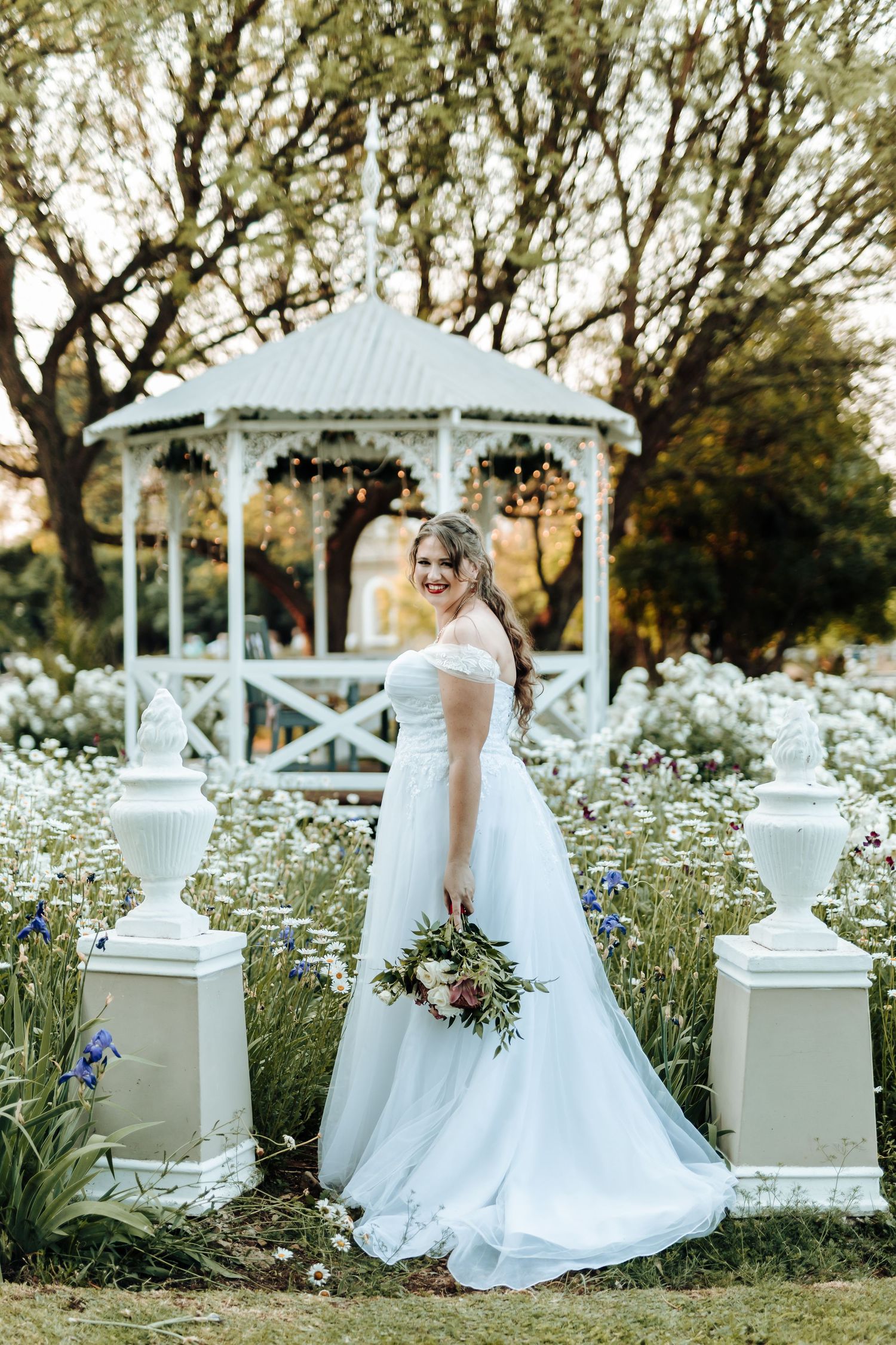 Bride in white gown poses by garden gazebo surrounded by white wildflowers.