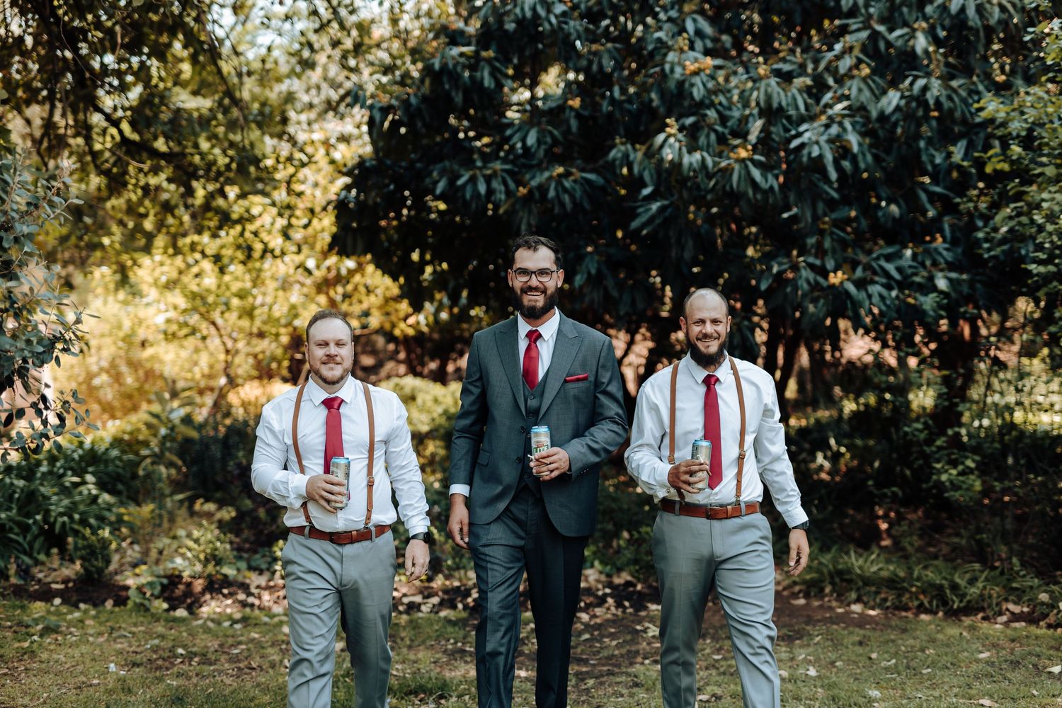 Wedding party in coordinated gray suits and red ties pose in garden setting.