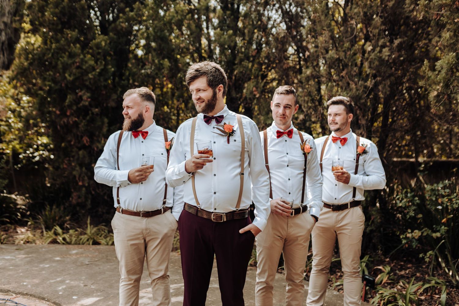 Wedding party in suspenders and bow ties hold drinks while posing outdoors.
