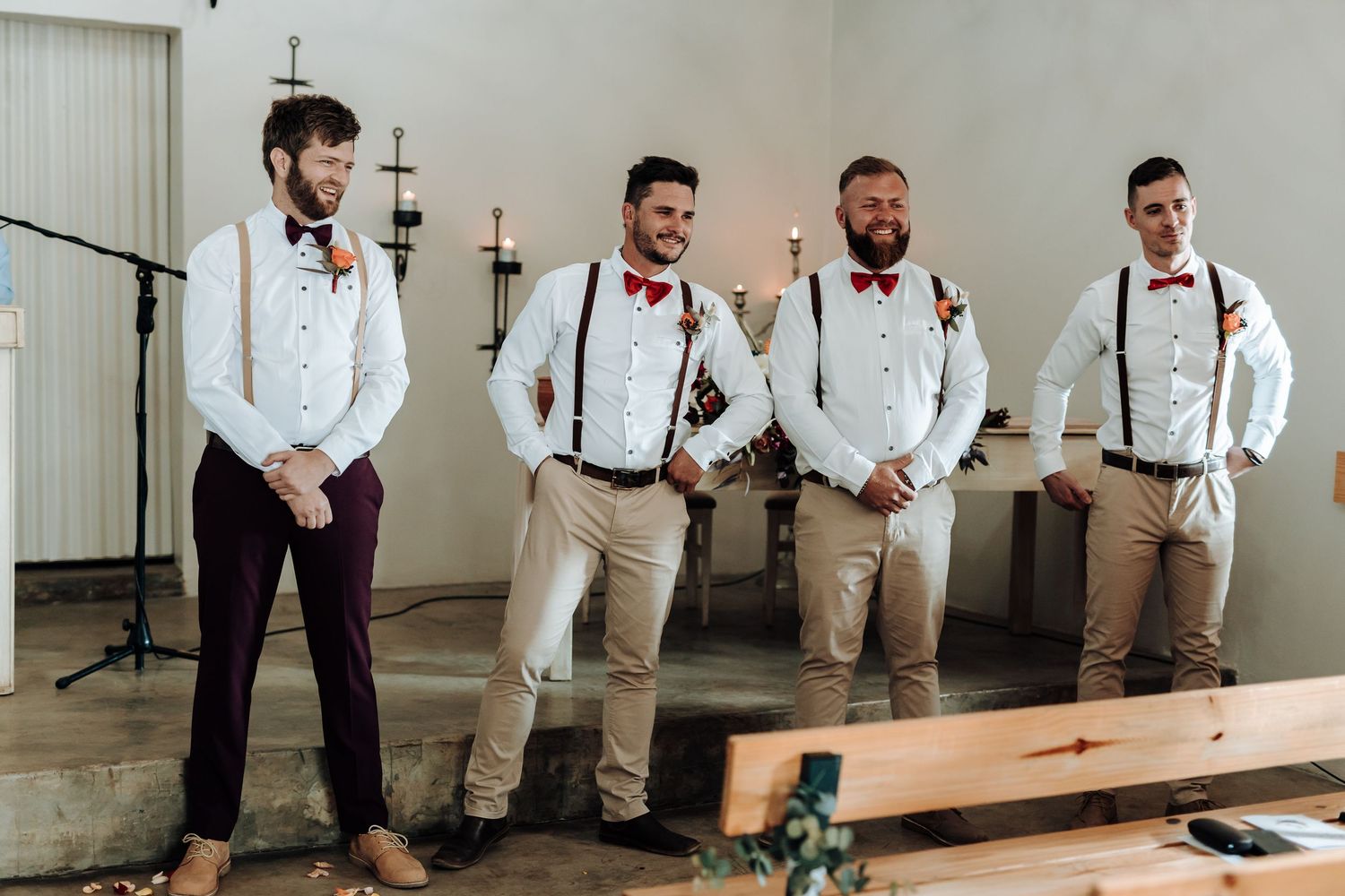 Groomsmen in casual wedding attire wearing suspenders and bowties stand together in a rustic venue.