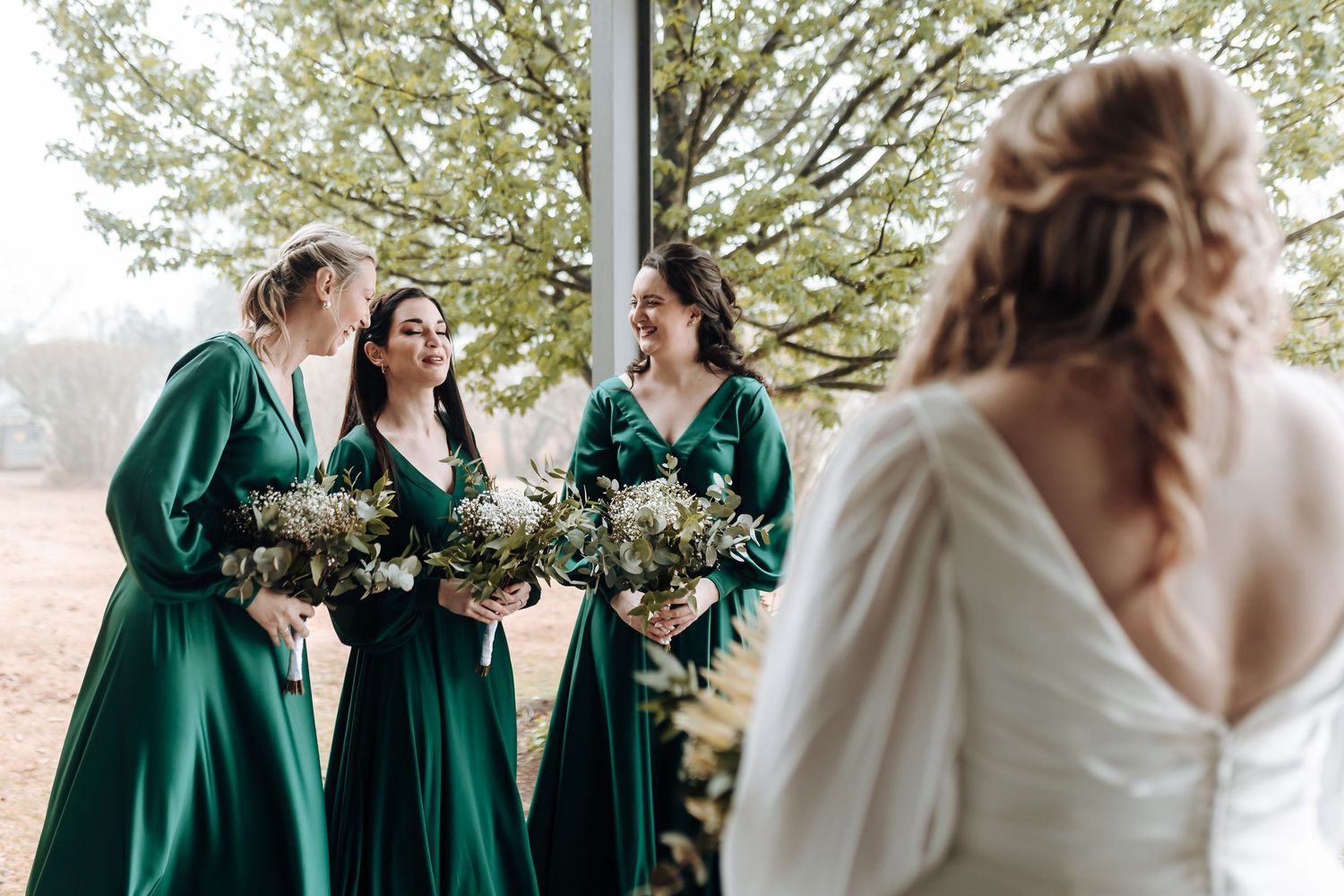 Bridesmaids wearing matching green dresses with bouquets talking to bride outdoors.