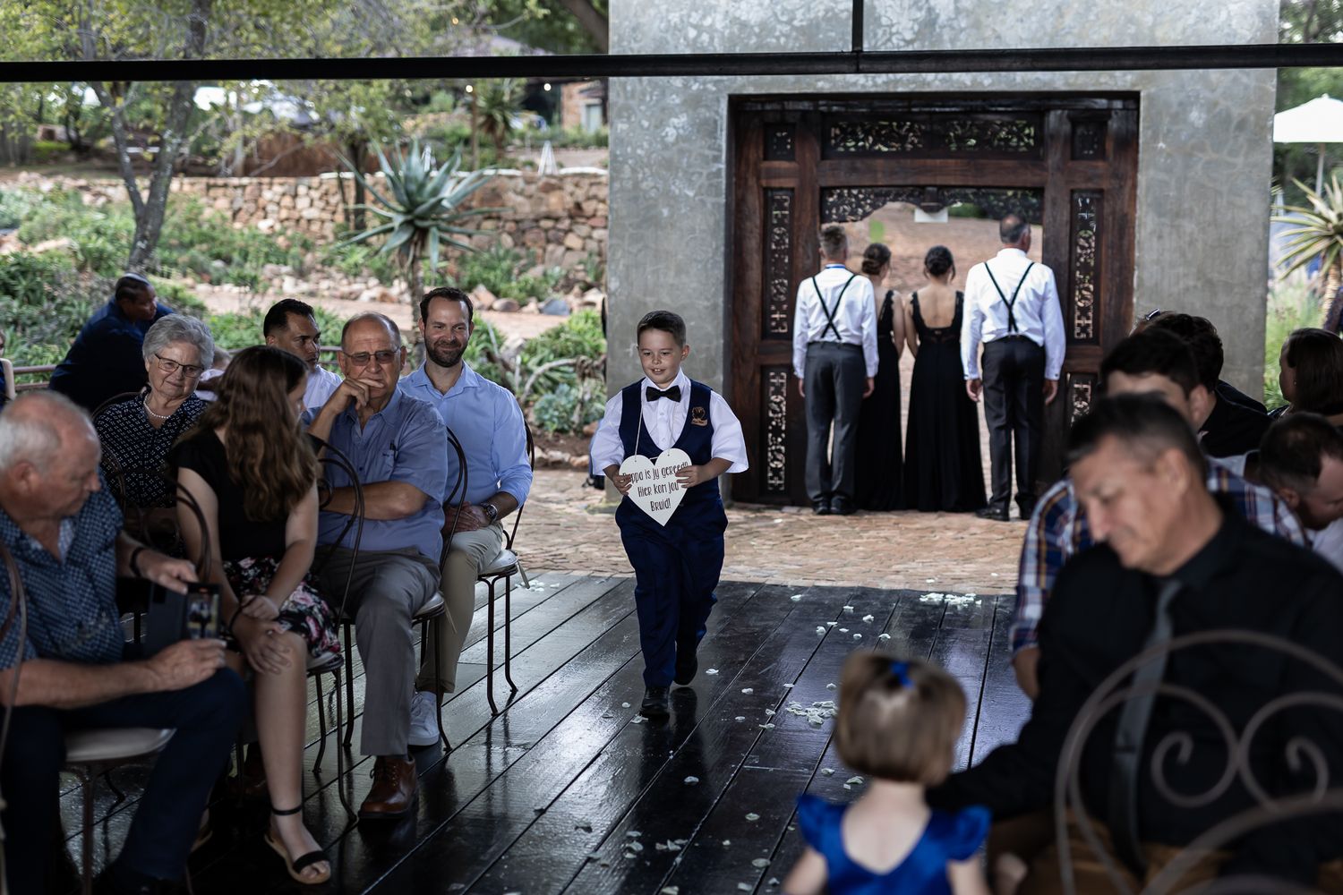 Wedding ceremony processional with bridal party members and seated guests on covered wooden deck.