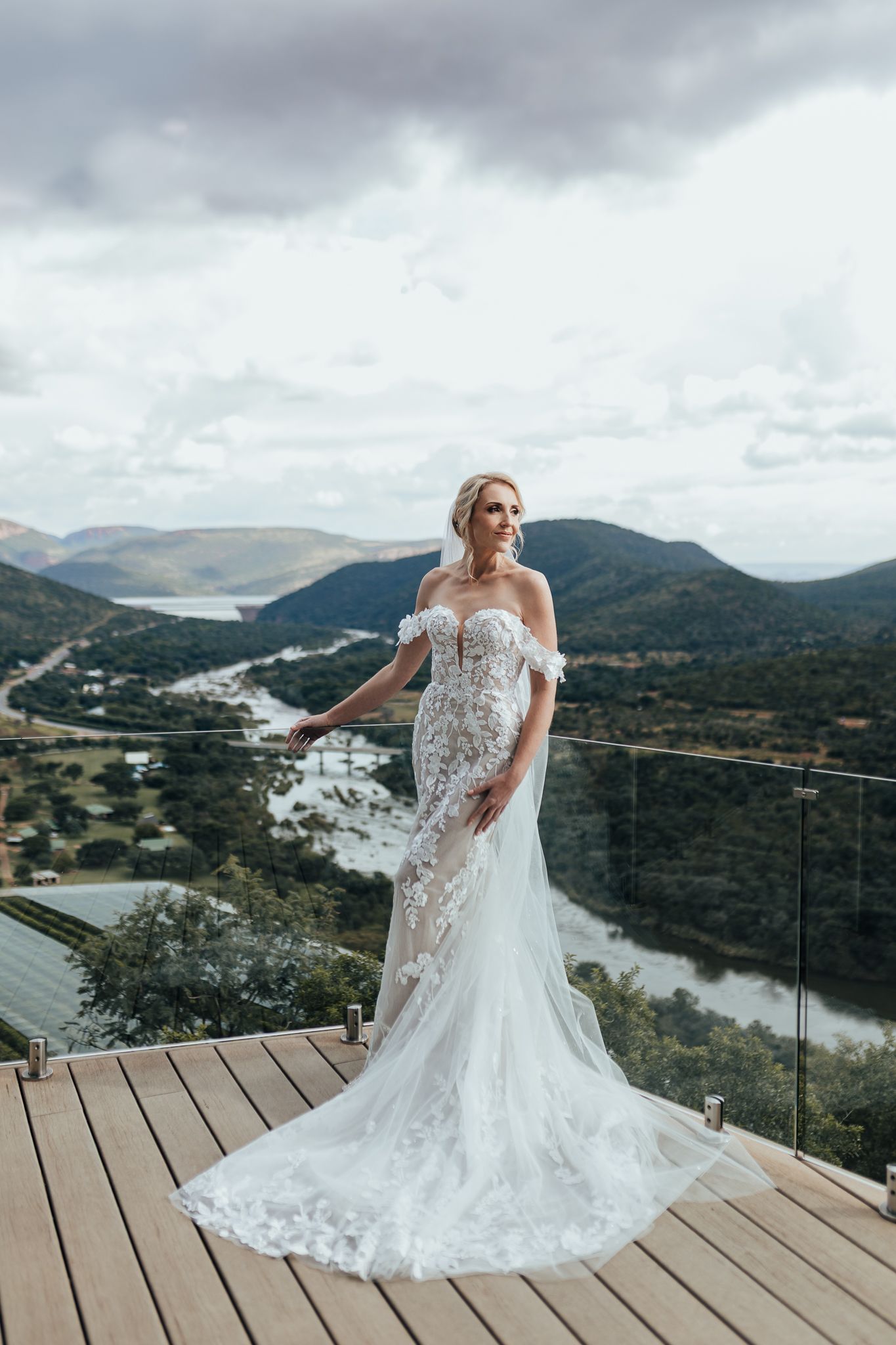 Bride in lace wedding dress poses on deck overlooking scenic mountain valley landscape.