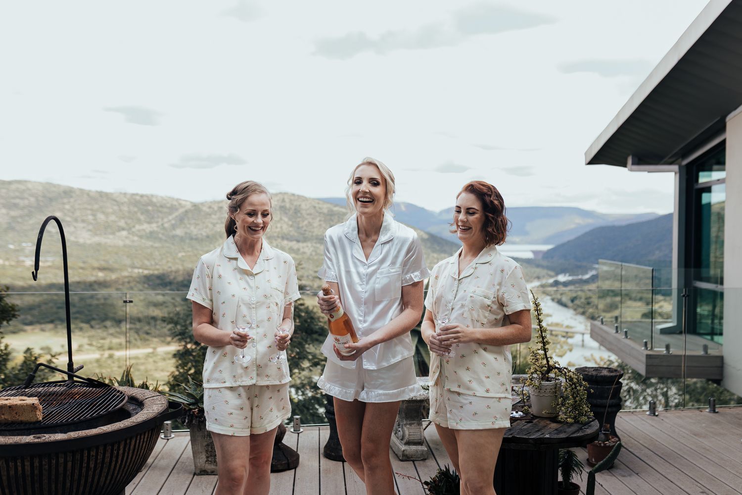 Three people in white outfits sharing drinks on outdoor deck with mountain views.
