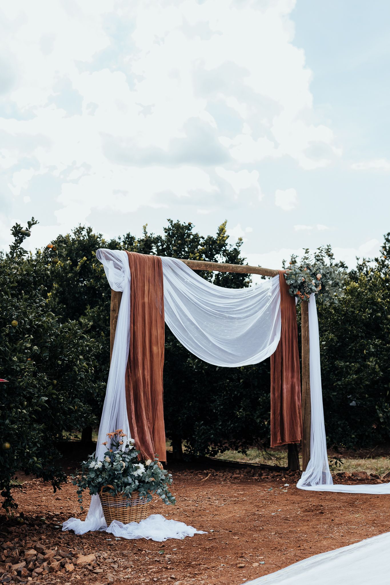 Wedding ceremony arch draped with white and terracotta fabric against garden backdrop.