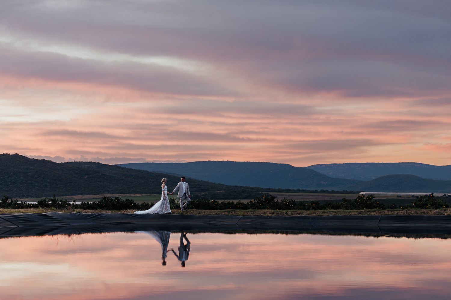Wedding couple reflected in water during pink sunset with mountain backdrop.