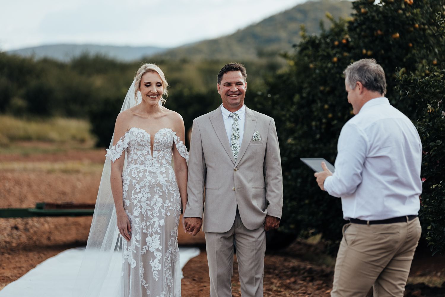 Bride in lace gown and groom in beige suit holding hands during outdoor ceremony.
