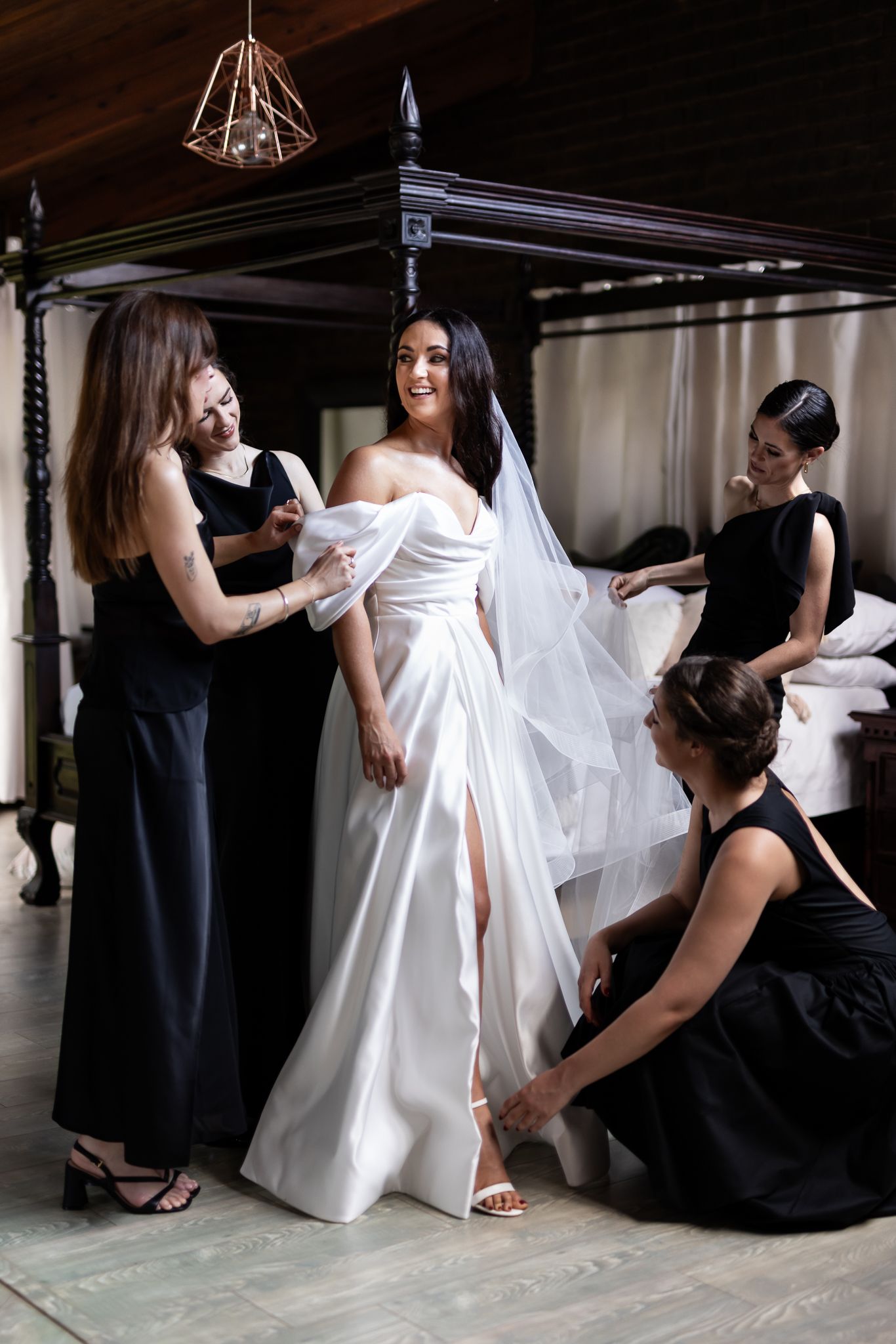 Bridesmaids help adjust a white wedding gown and veil during bridal preparations in an elegant room with canopy bed.
