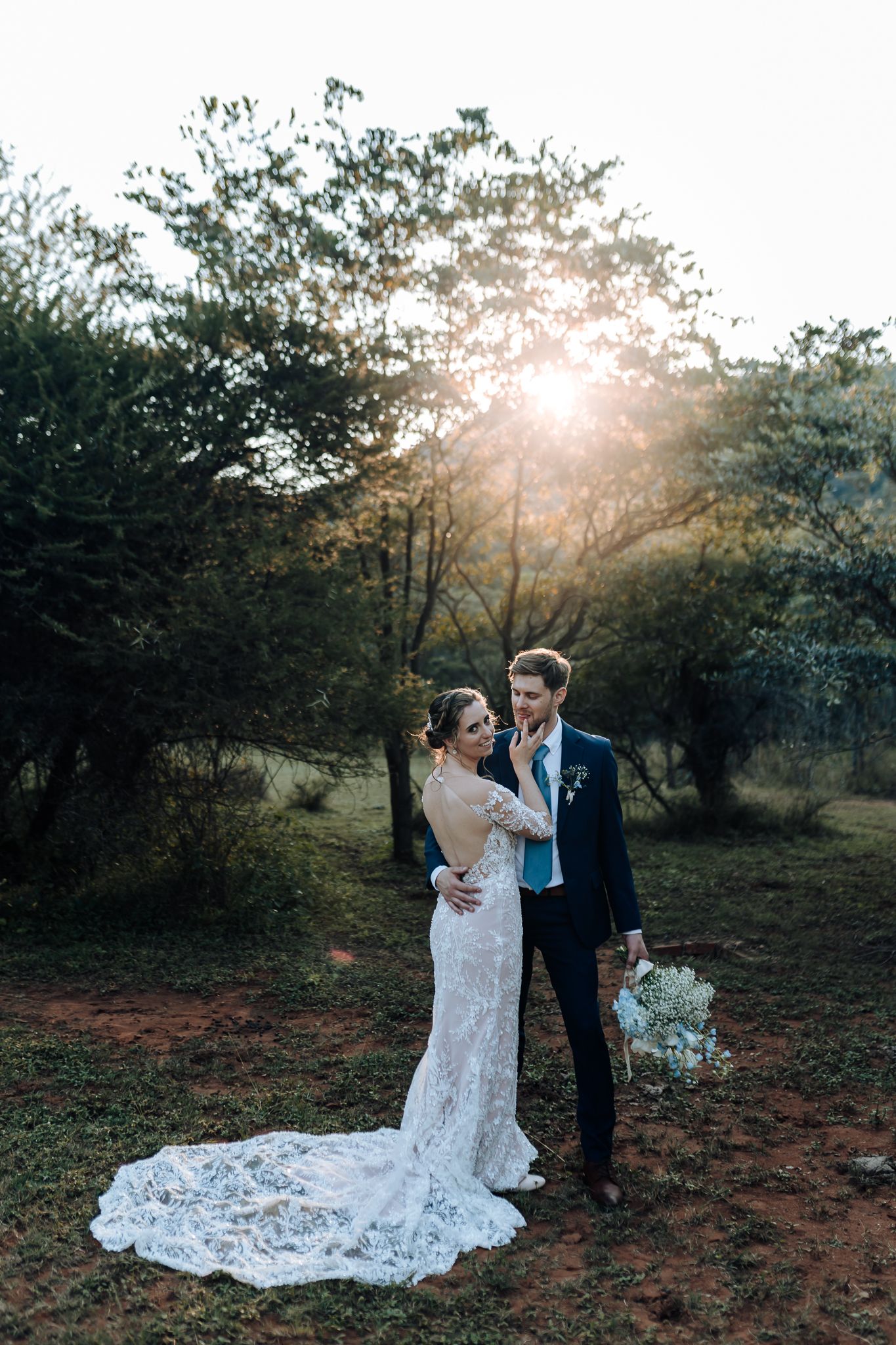 Stunning wedding photo with dramatic sunburst through trees and bride's lace train extending along ground.