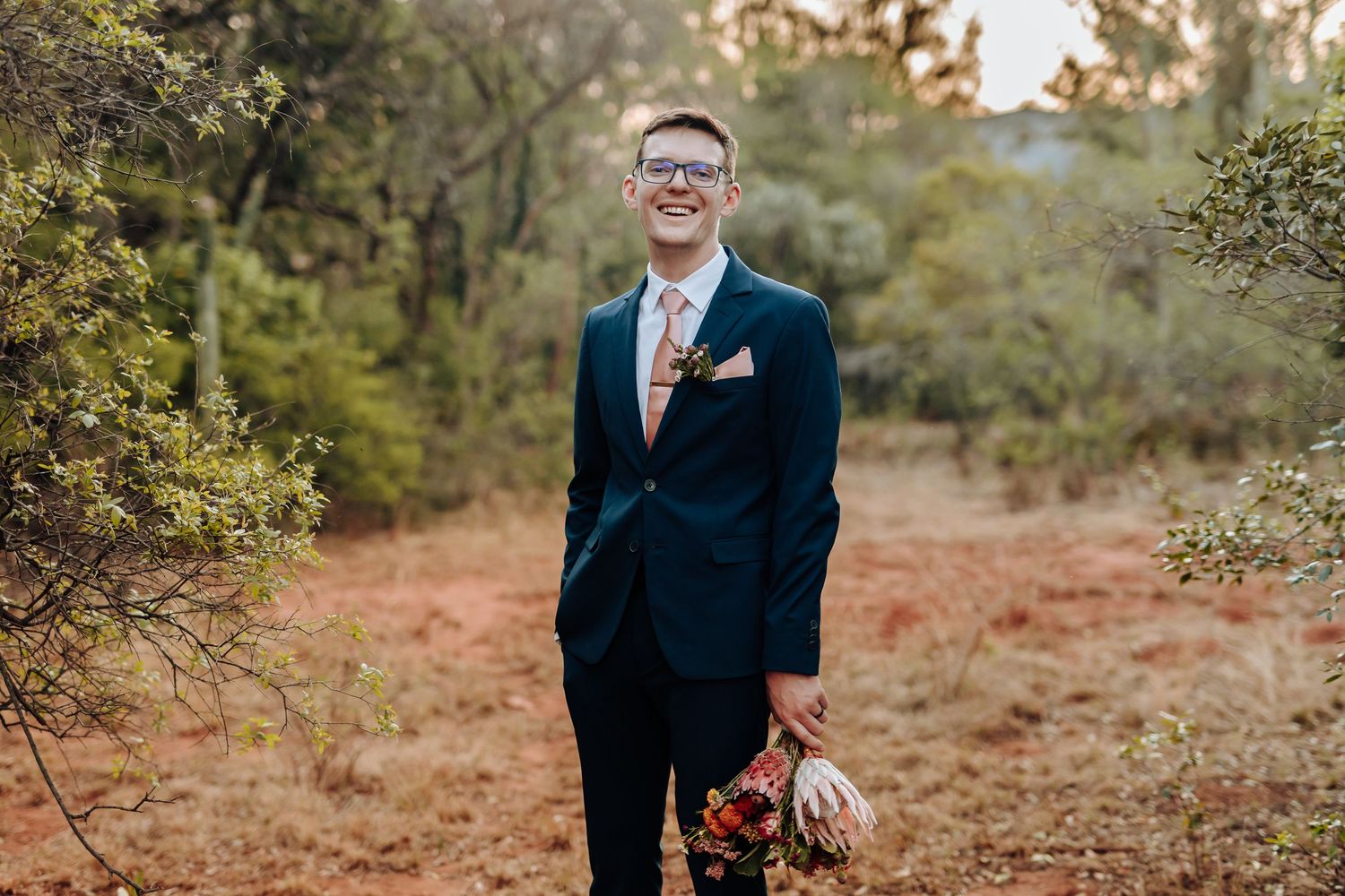 A groom in a navy suit poses in an autumn forest setting with warm lighting and fall foliage in the background.