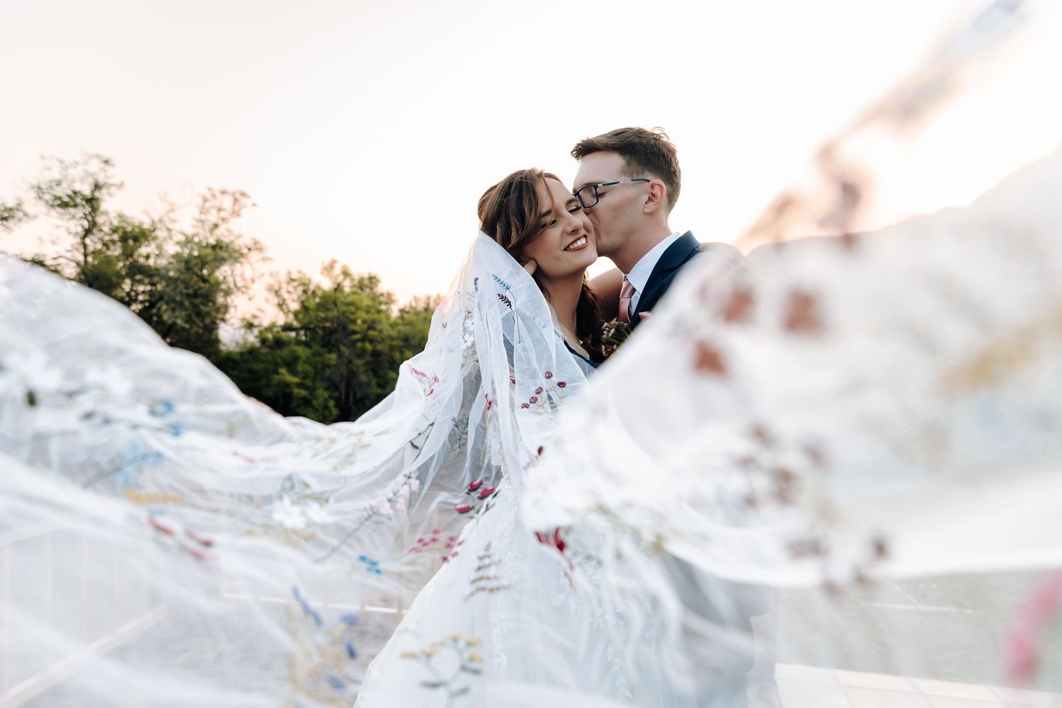 A couple in wedding attire shares a romantic moment under flowing white fabric during an outdoor photo session.