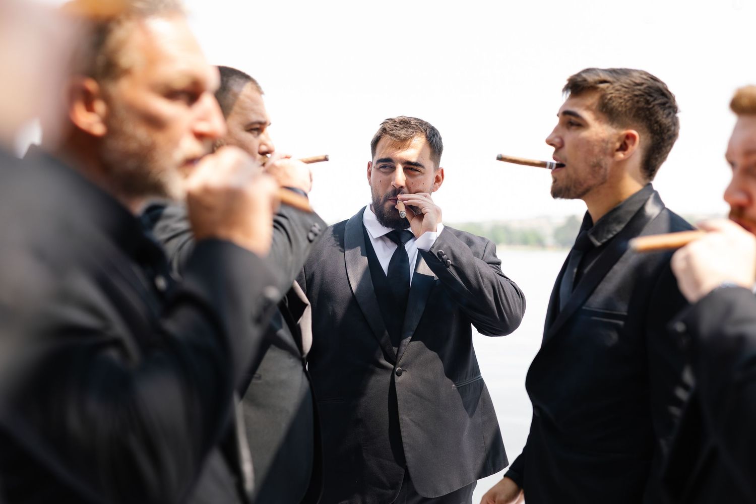 A group of men in dark suits standing together while smoking cigars outdoors.