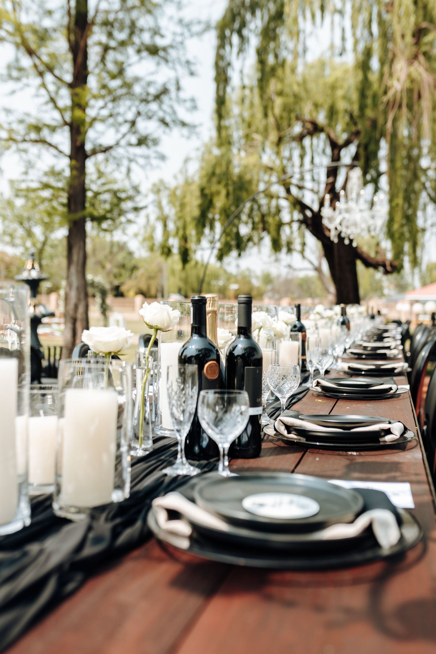 Elegant table setting with black plates and blue glass bottles arranged on dark wood surface against outdoor background.