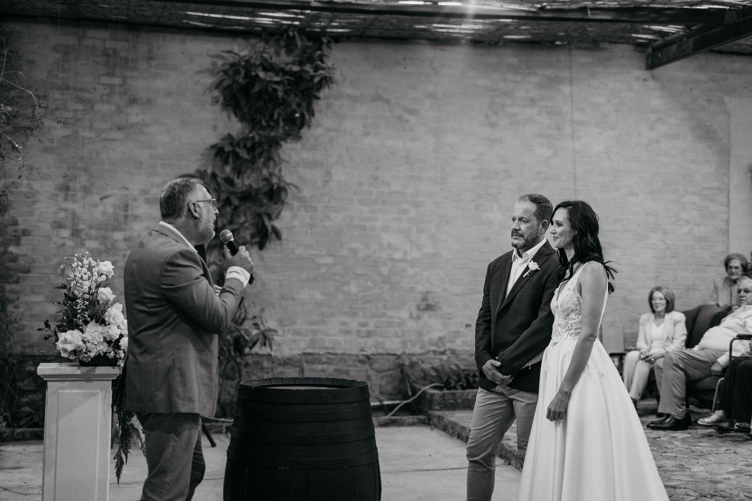 Couple stands at wedding altar as officiant speaks during outdoor brick wall ceremony with seated guests in background.