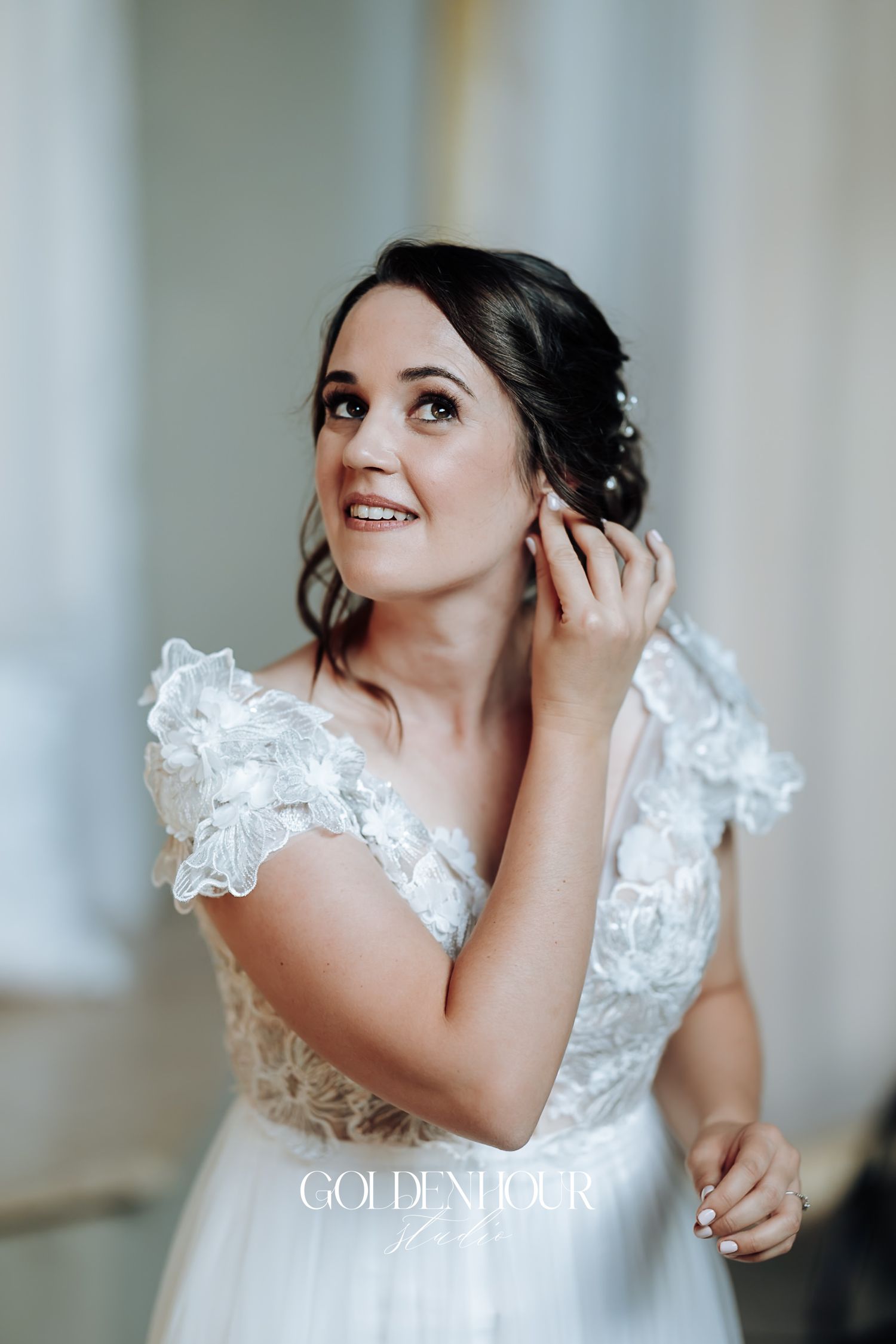 Bride in a lace wedding dress adjusting her earring while looking up in soft natural light.