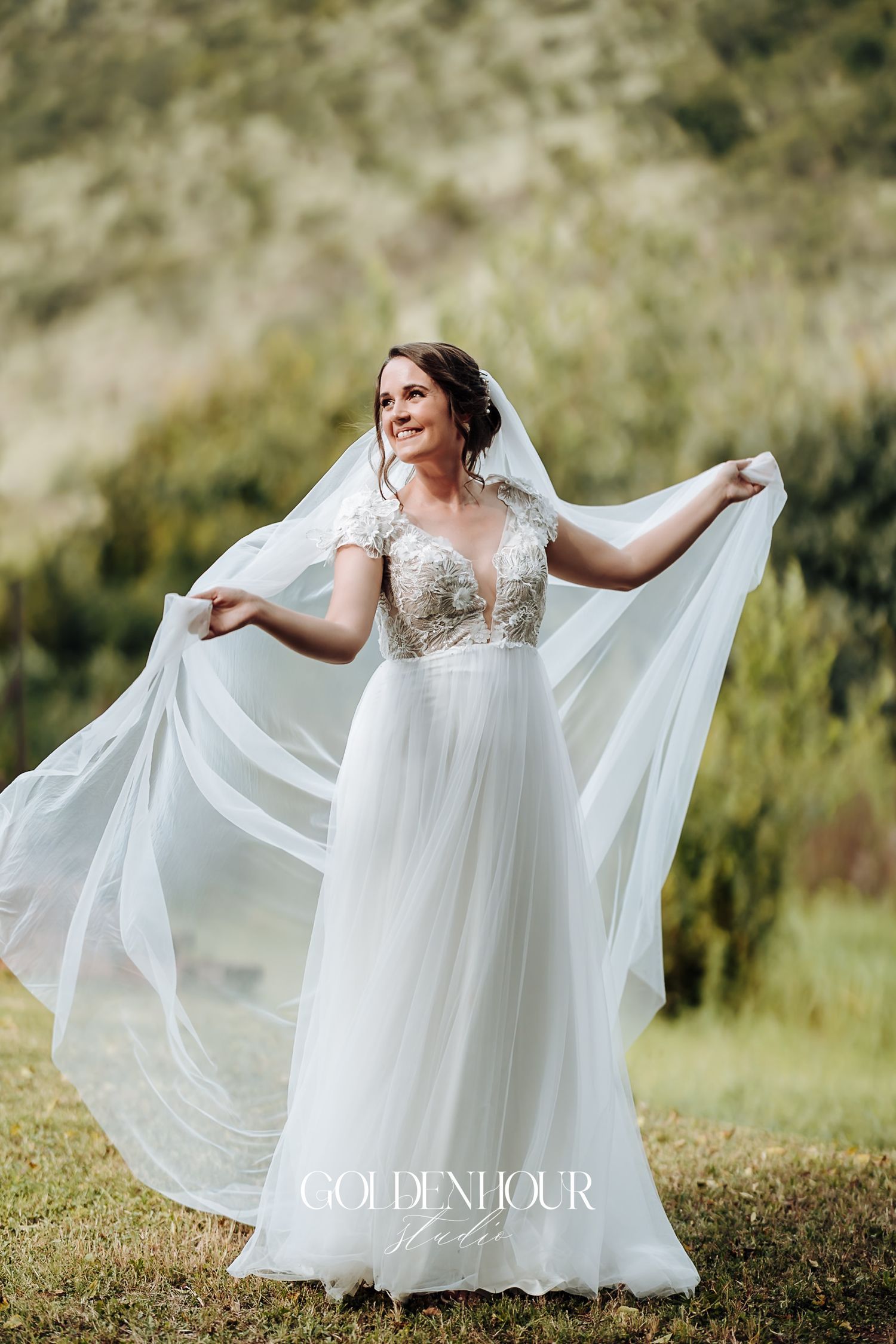 Bride in lace and tulle gown spreads her veil wide in a scenic outdoor wedding portrait.