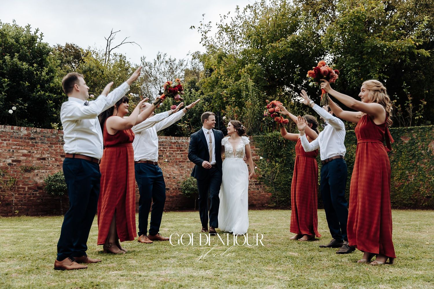 Wedding party in red dresses and navy suits celebrate with bouquets raised in garden setting.