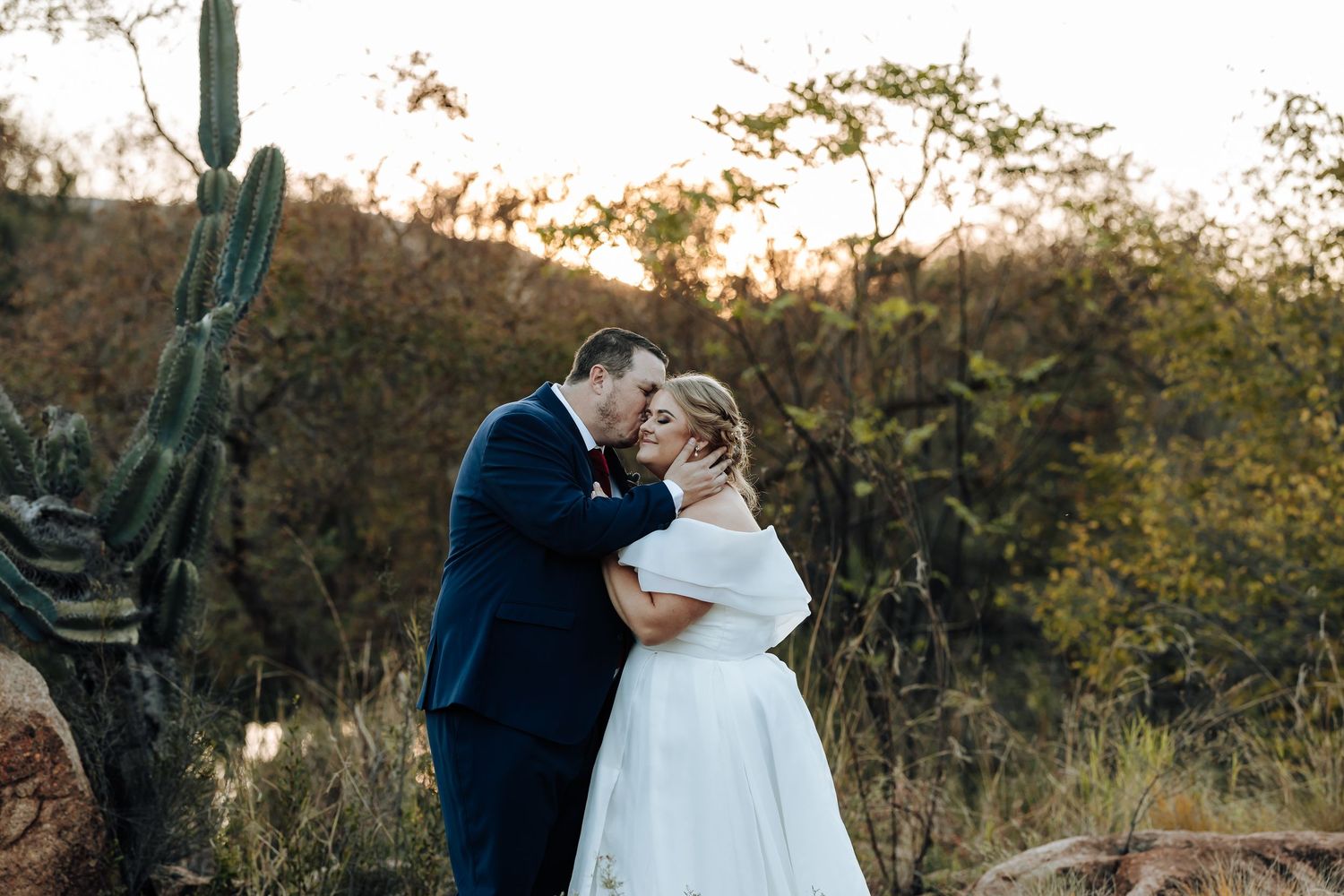 Bride and groom share a romantic embrace at sunset in a desert landscape with saguaro cactus and golden foliage.