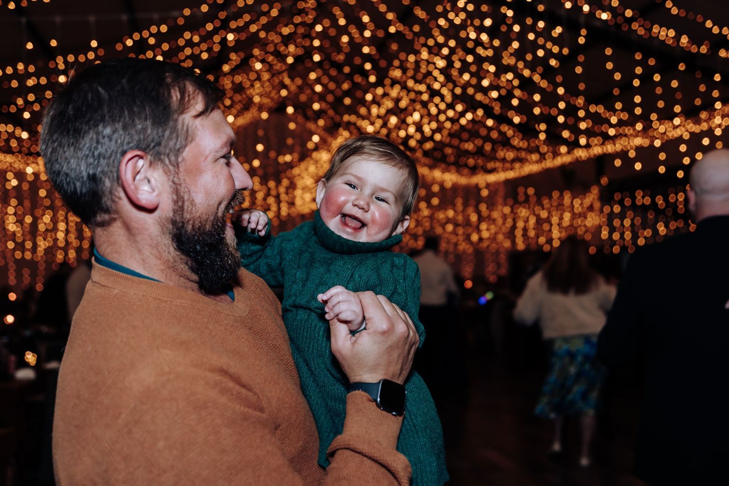 A bearded man in an orange sweater joyfully holds a laughing toddler in green amid warm fairy lights.
