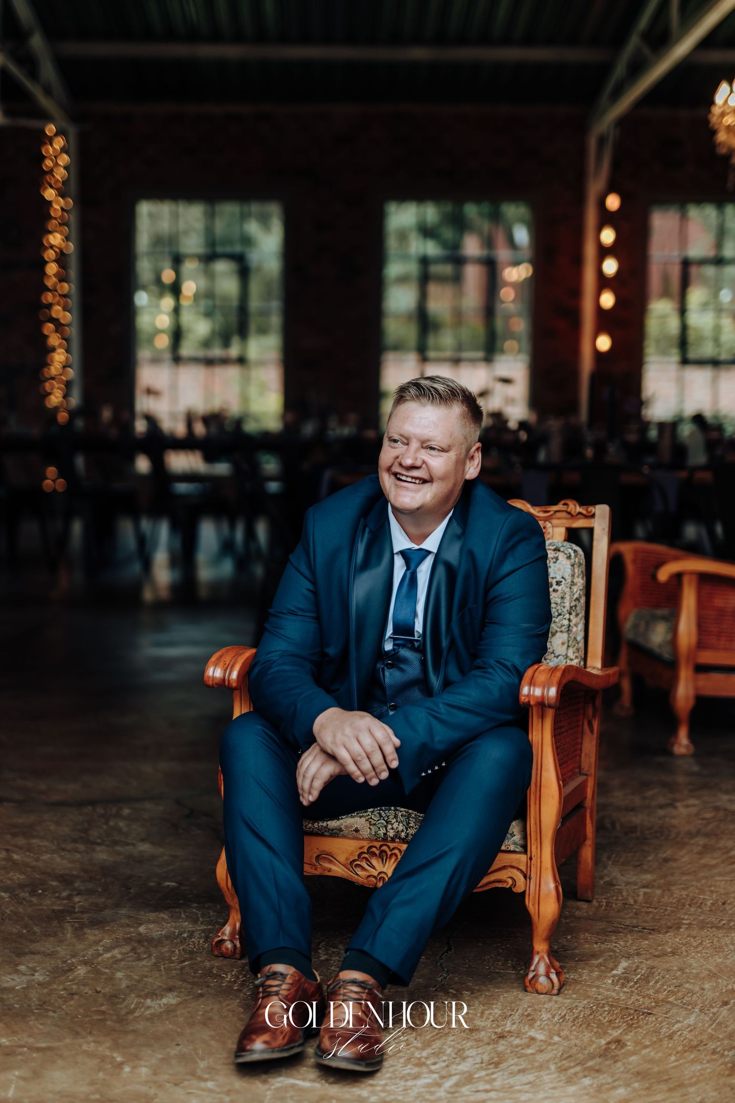 Smiling groom in navy blue suit sitting in ornate chair at elegant wedding reception venue.
