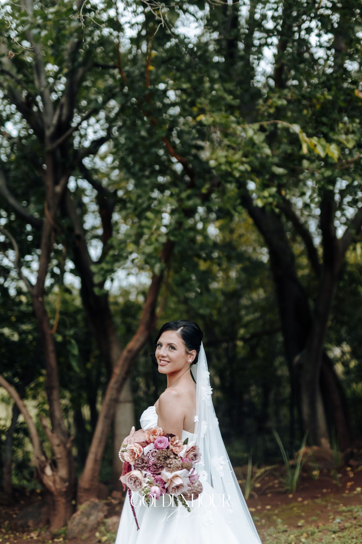 Smiling bride in white gown holds pink bouquet outdoors among lush green trees.