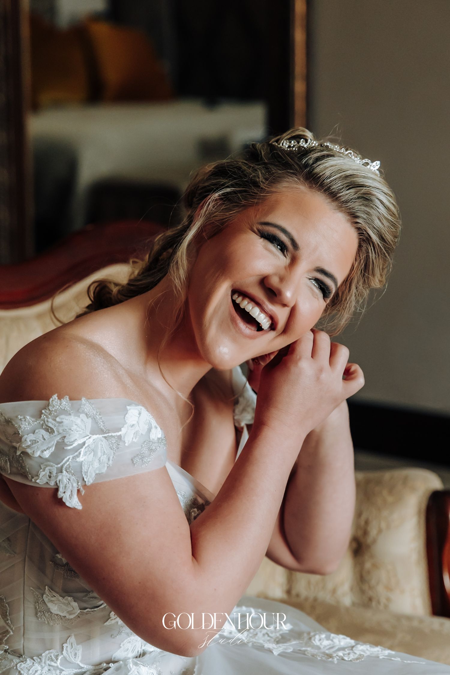 Joyful bride in an off-shoulder lace wedding gown and tiara laughing while seated on an elegant sofa.