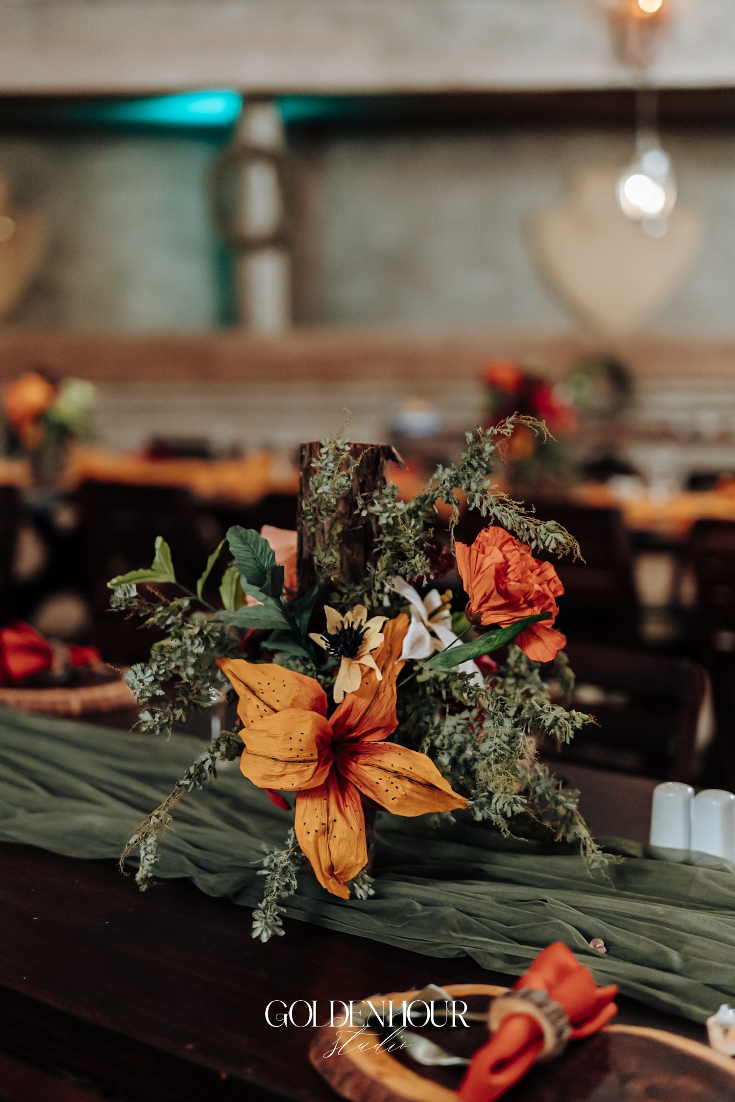 Rustic wedding table centerpiece with orange lilies, green foliage, and red blooms on a dark wood table.