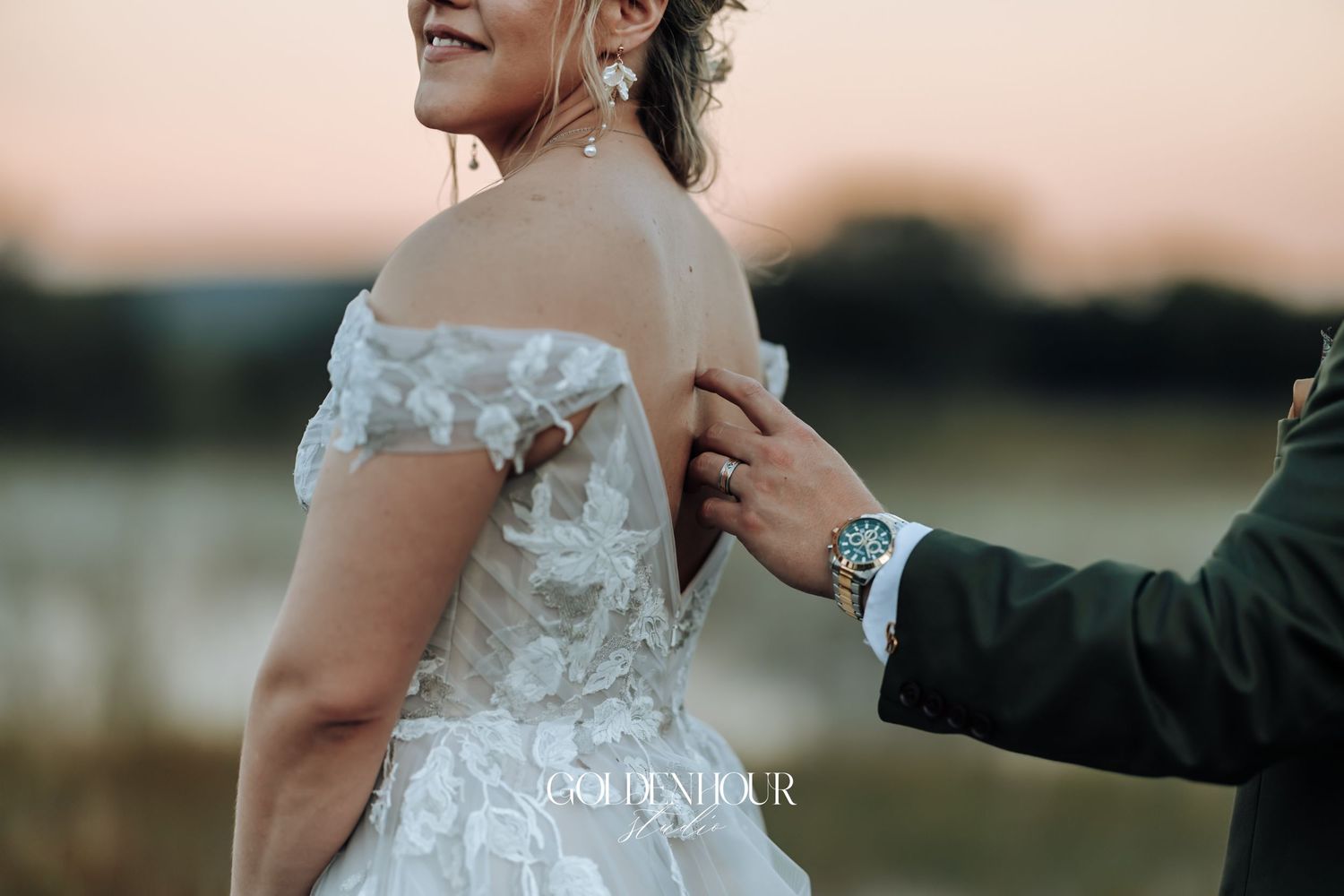 Groom gently touching the back of bride's intricate lace off-shoulder wedding gown during a sunset portrait session.