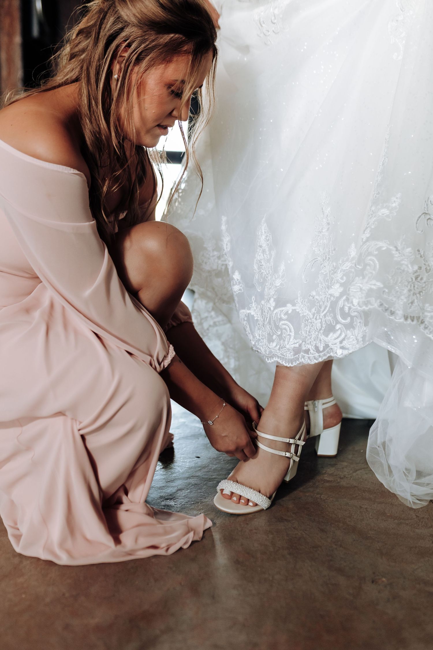 Someone in a pink dress helps adjust white wedding shoes on a bride wearing a lacy white gown.