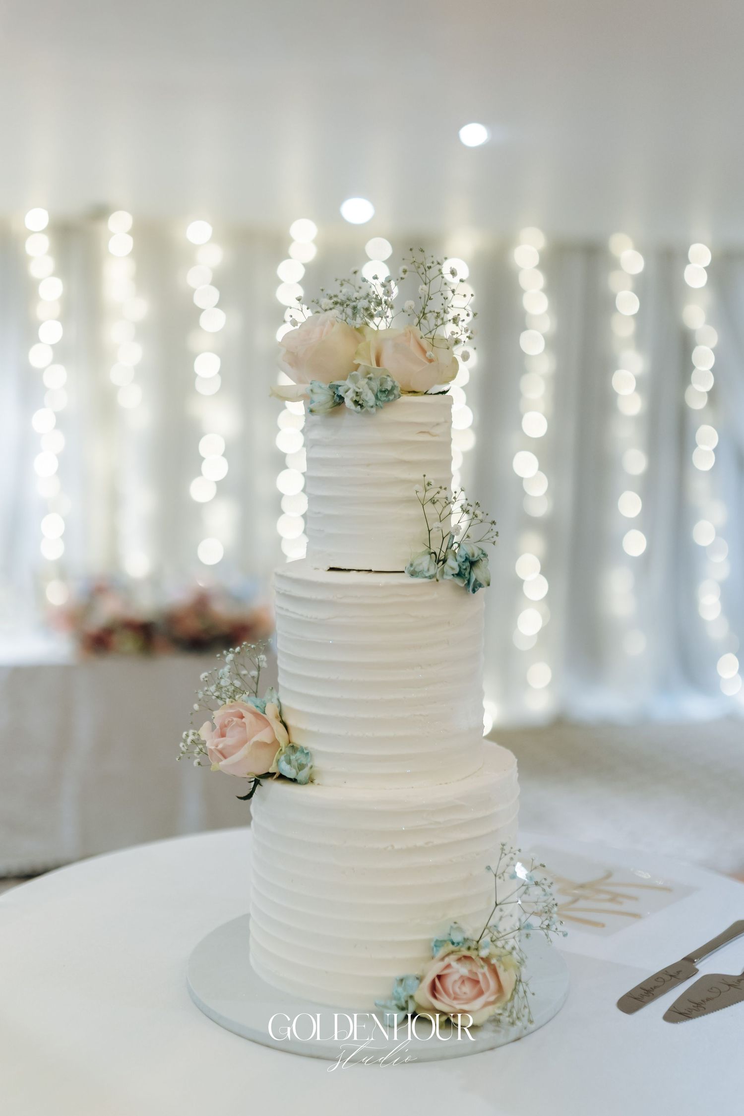 Three-tiered white wedding cake with textured frosting and blush roses against a backdrop of string lights.