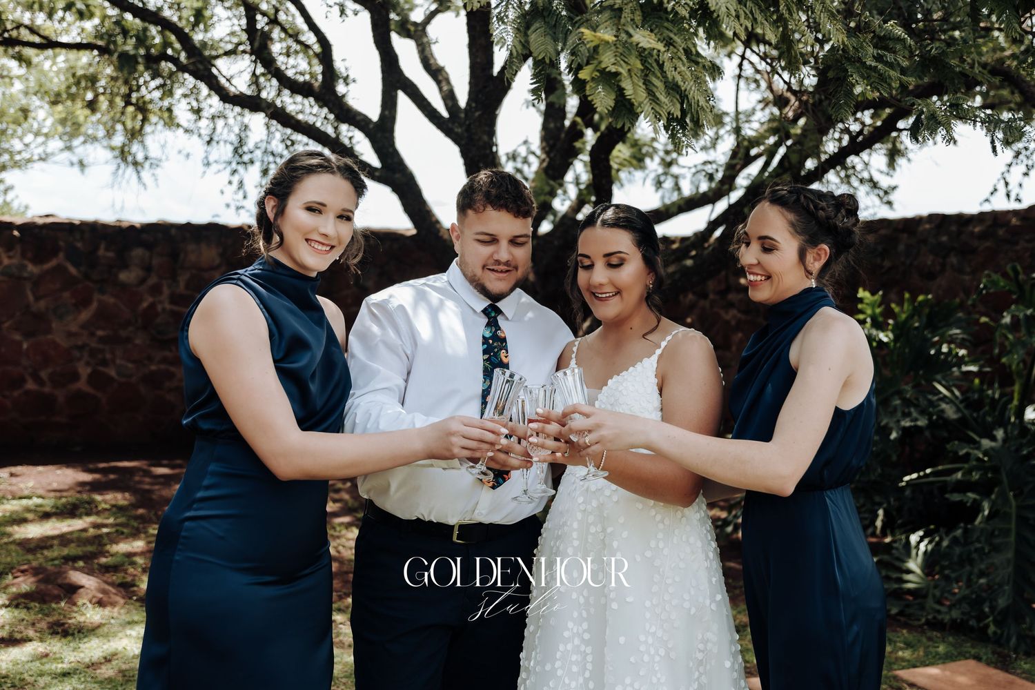 Wedding party members in navy blue and white attire share a celebratory toast outdoors under trees.