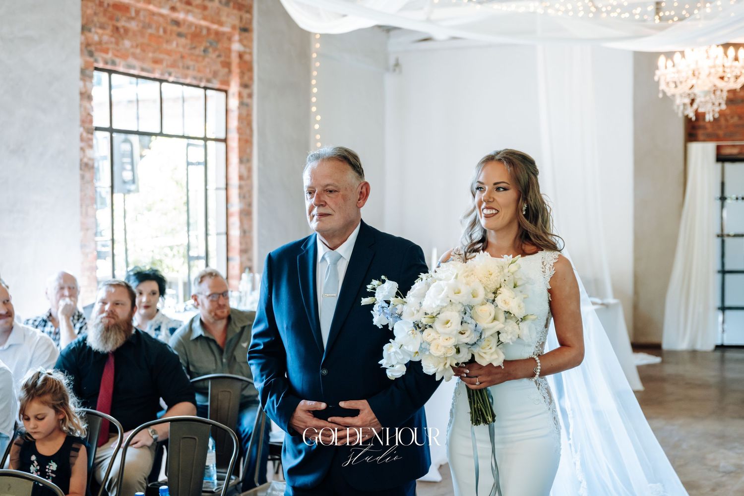 Elegant wedding ceremony in a bright industrial venue with white floral arrangements and crystal chandeliers.
