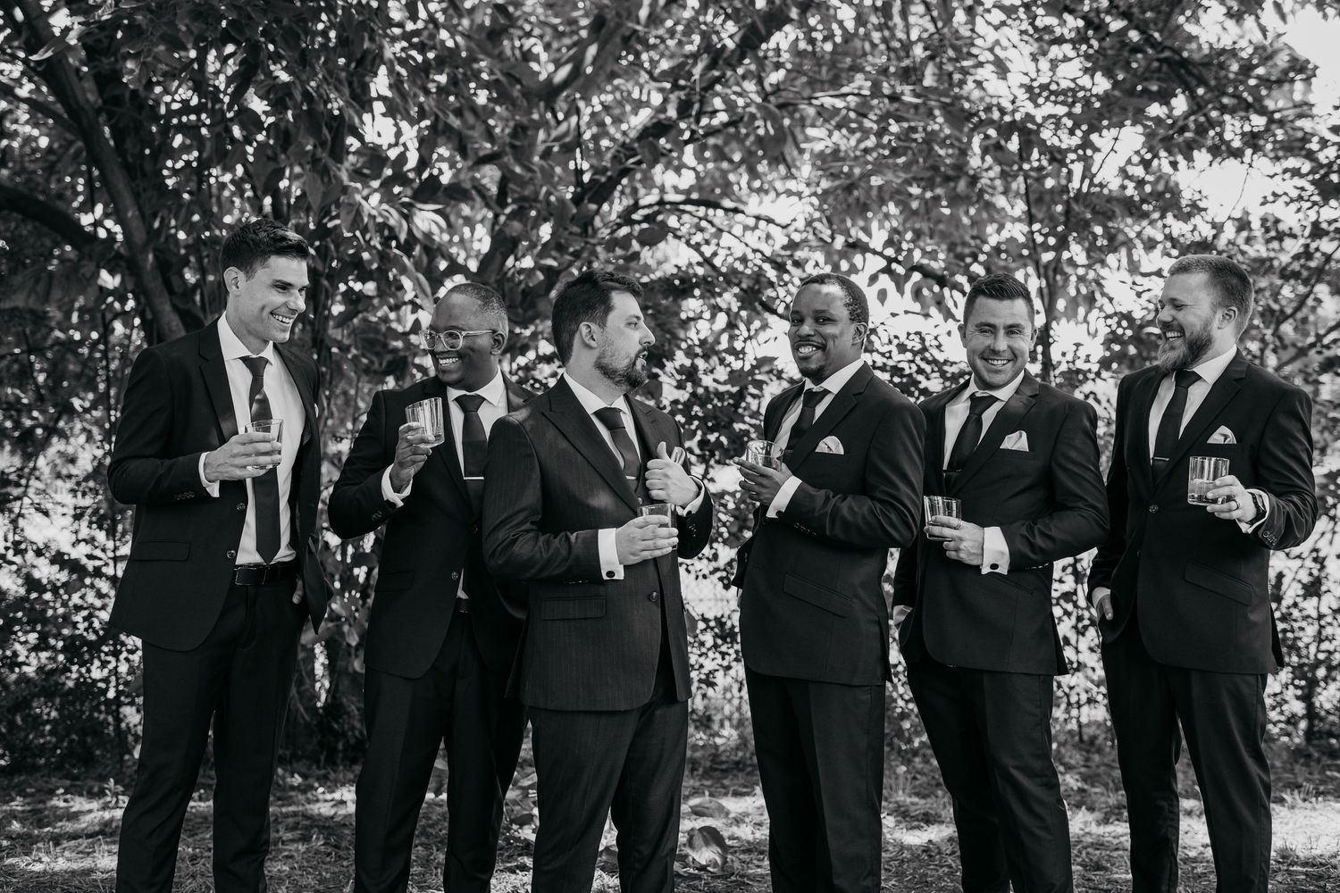 Black and white photo of groomsmen raising glasses in a celebratory toast under trees.