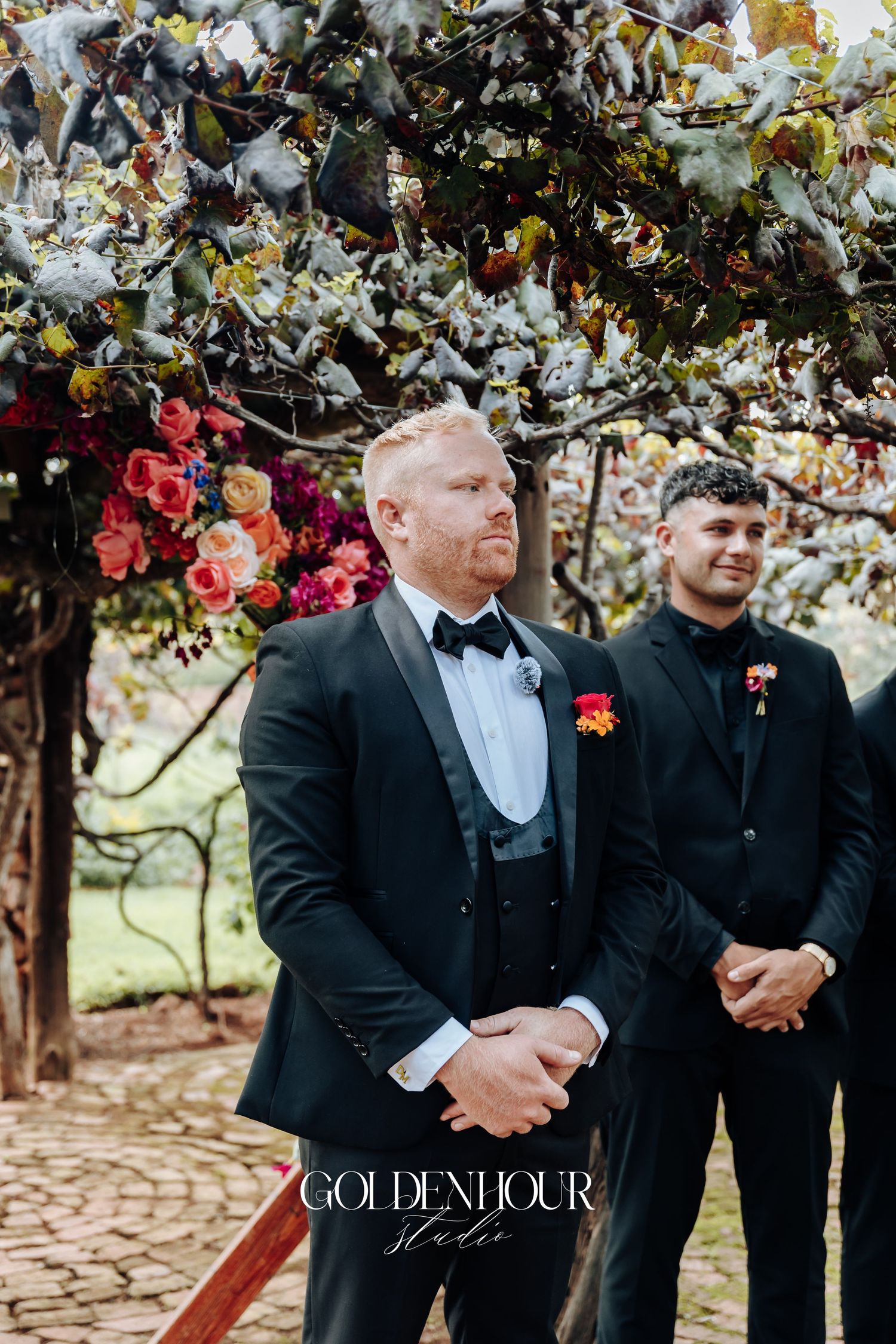 Groomsmen in black tuxedos and suits stand together outdoors under flowering trees during a wedding ceremony.