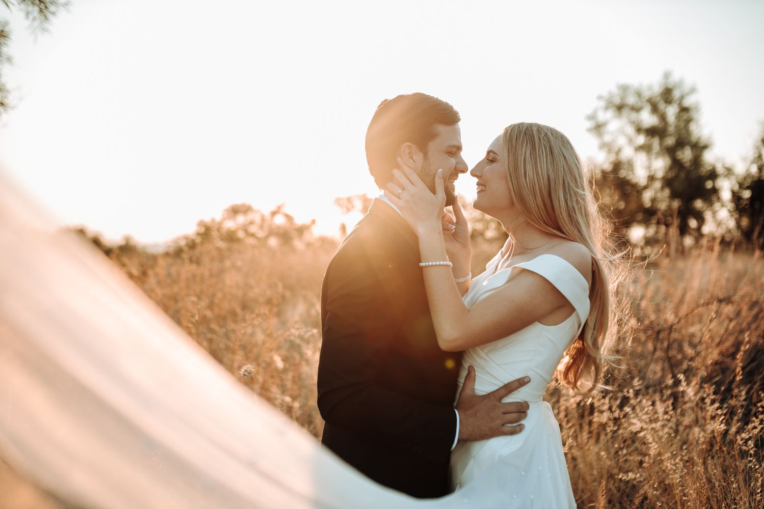 Romantic sunset wedding photo of couple in a golden field with flowing veil.