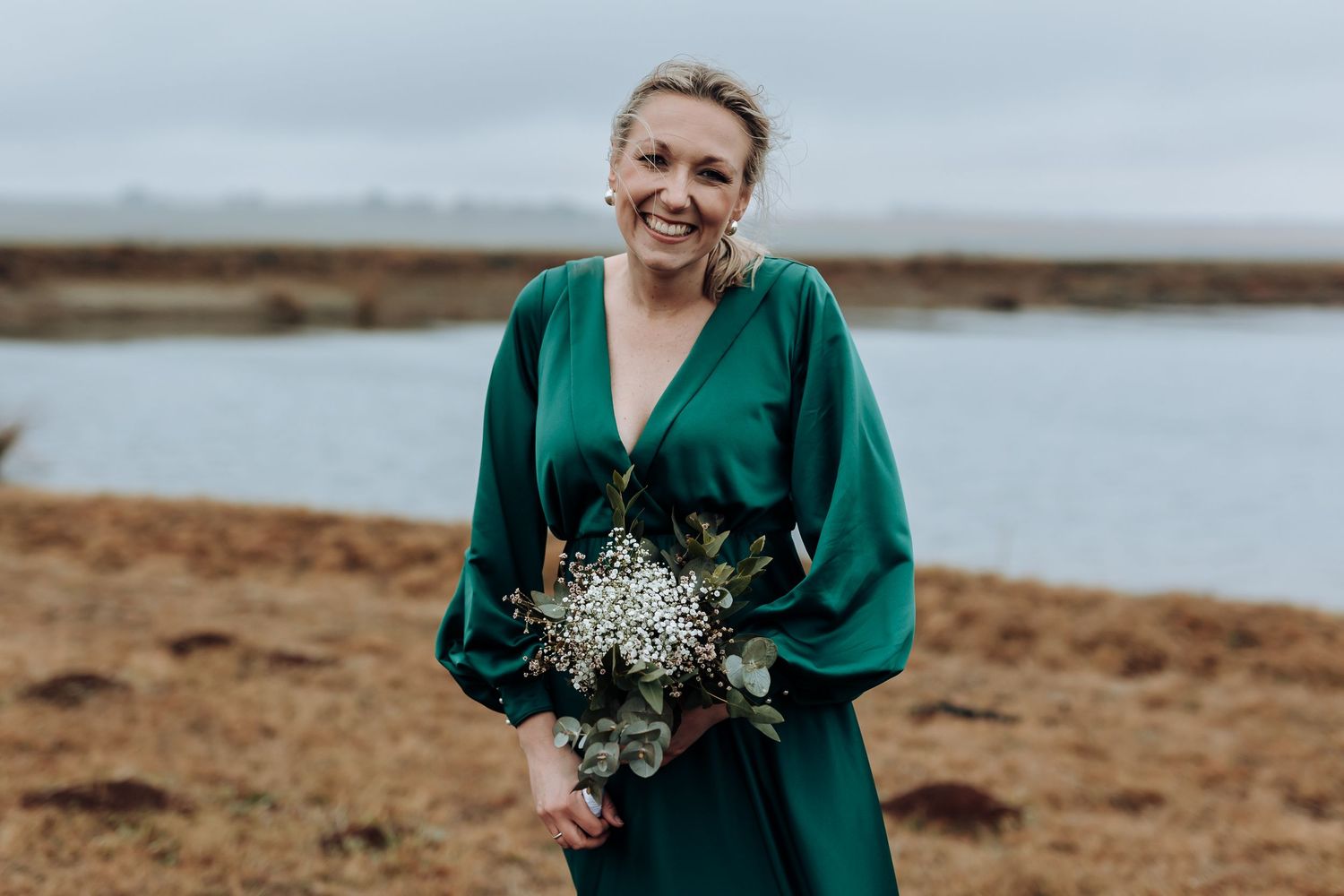 Bridesmaid in emerald green dress smiles while holding floral bouquet near water.