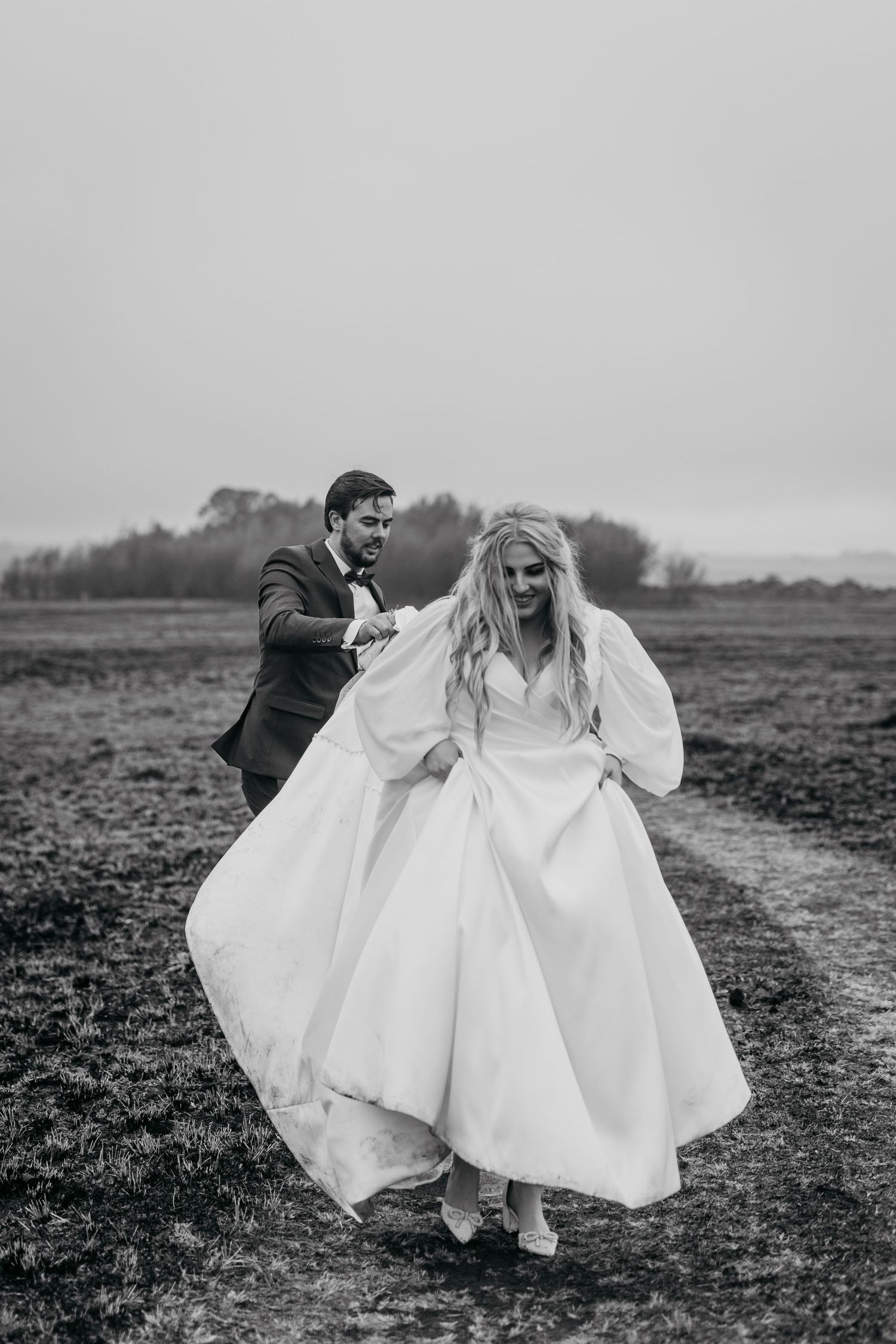 A windswept wedding couple runs playfully through a field on an overcast day.