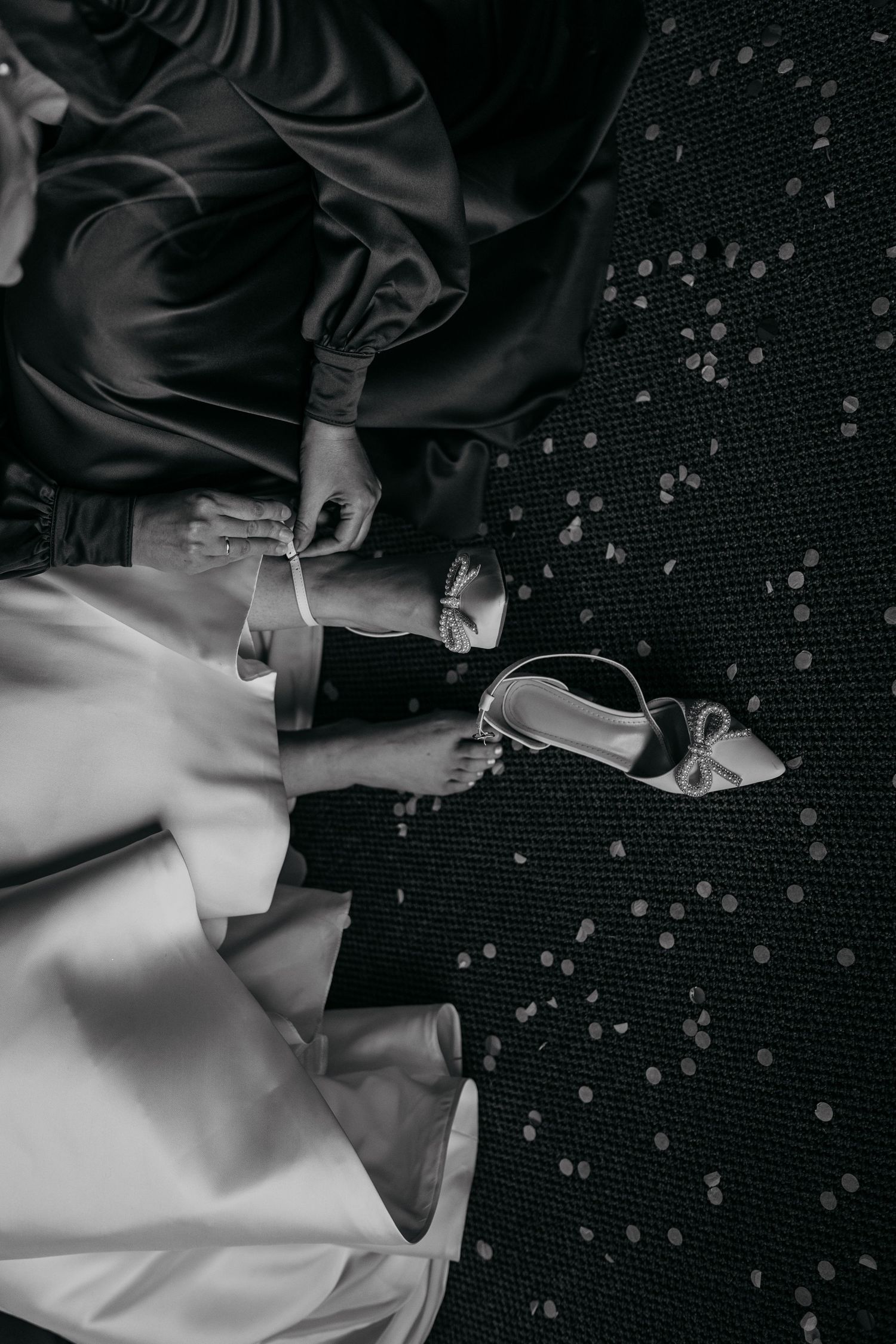 Wedding shoes and dress details in black and white with scattered confetti on dark carpet.