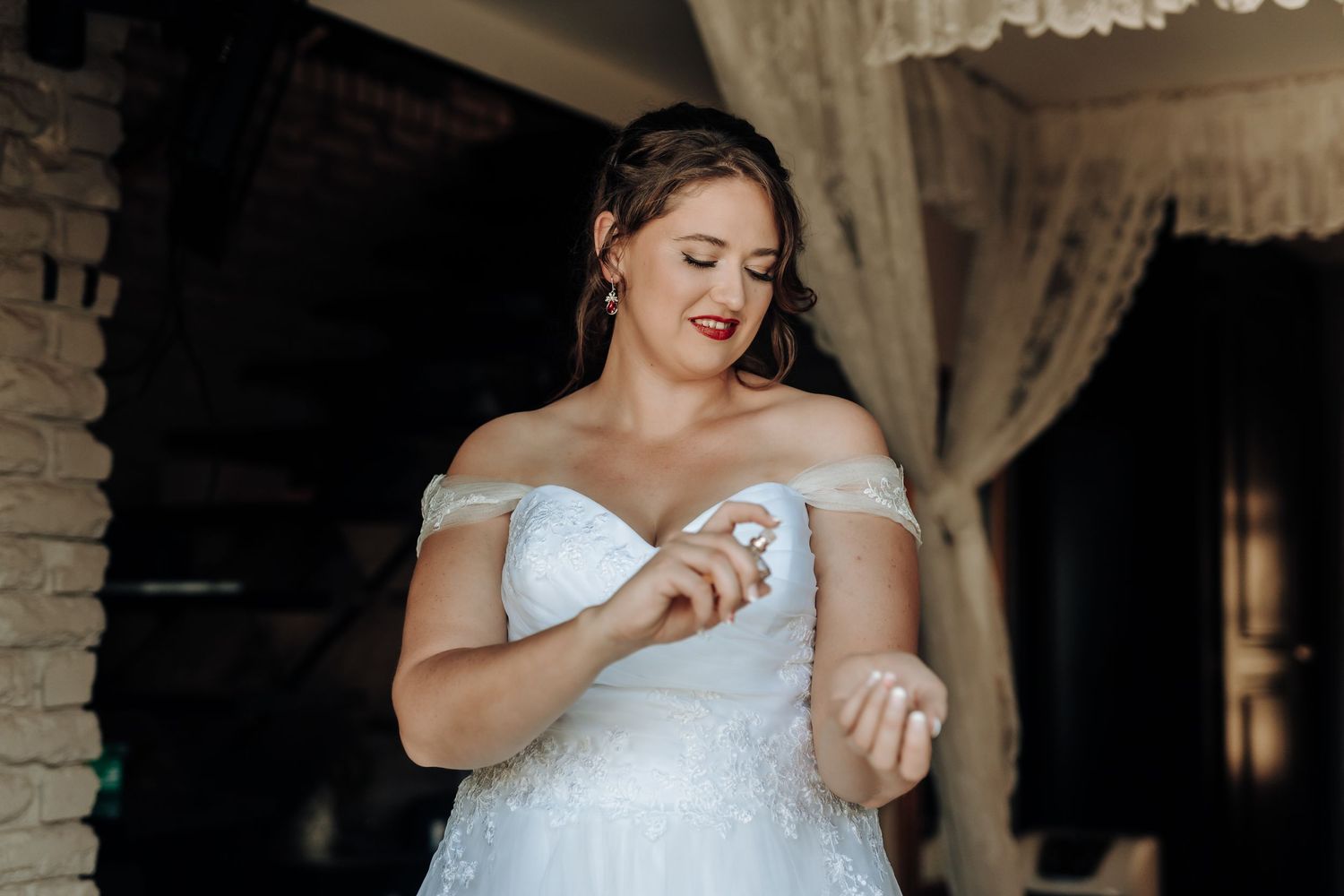 Bride in an off-shoulder lace gown applies perfume during wedding preparations.