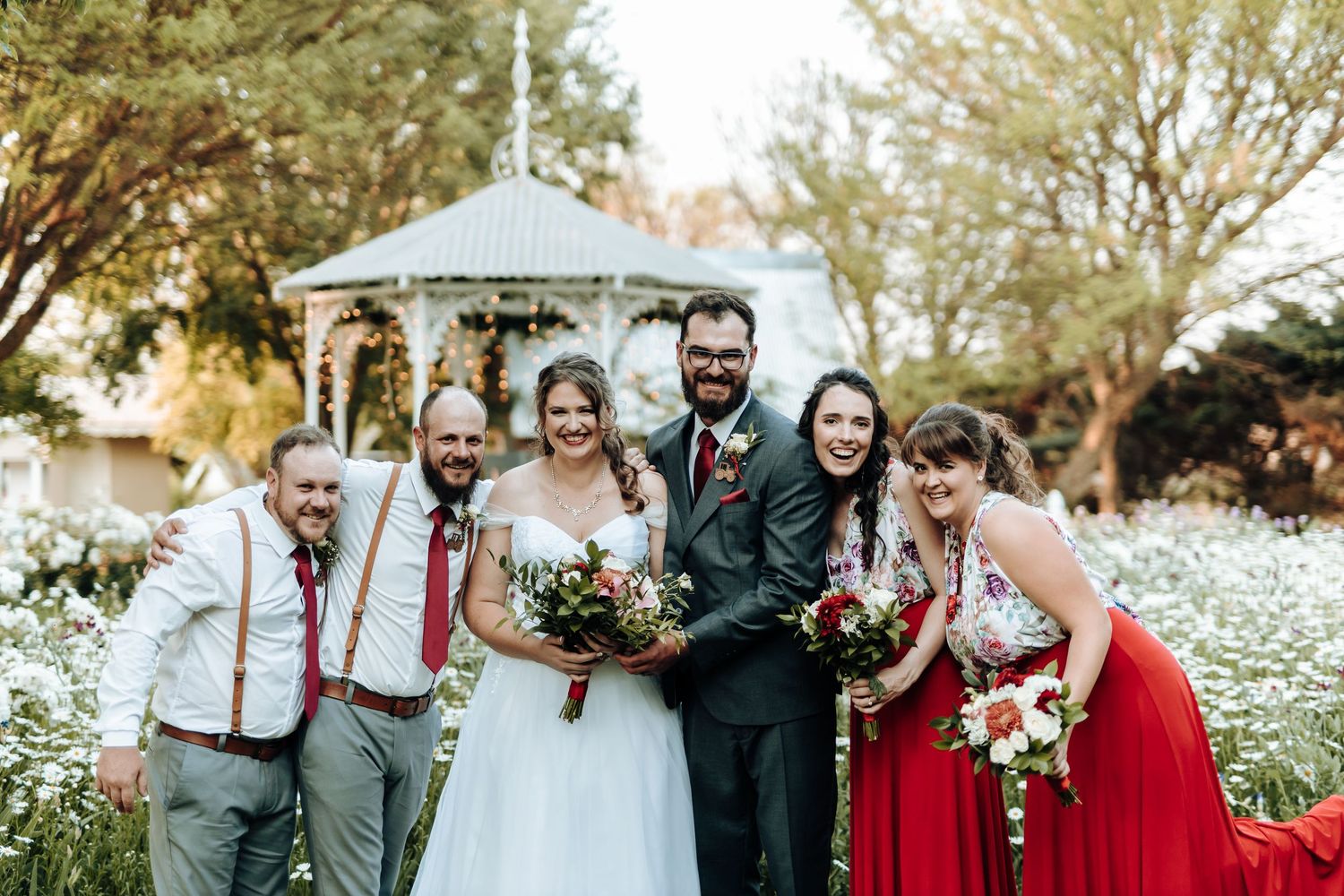 Wedding party gathers under garden gazebo surrounded by white wildflowers.