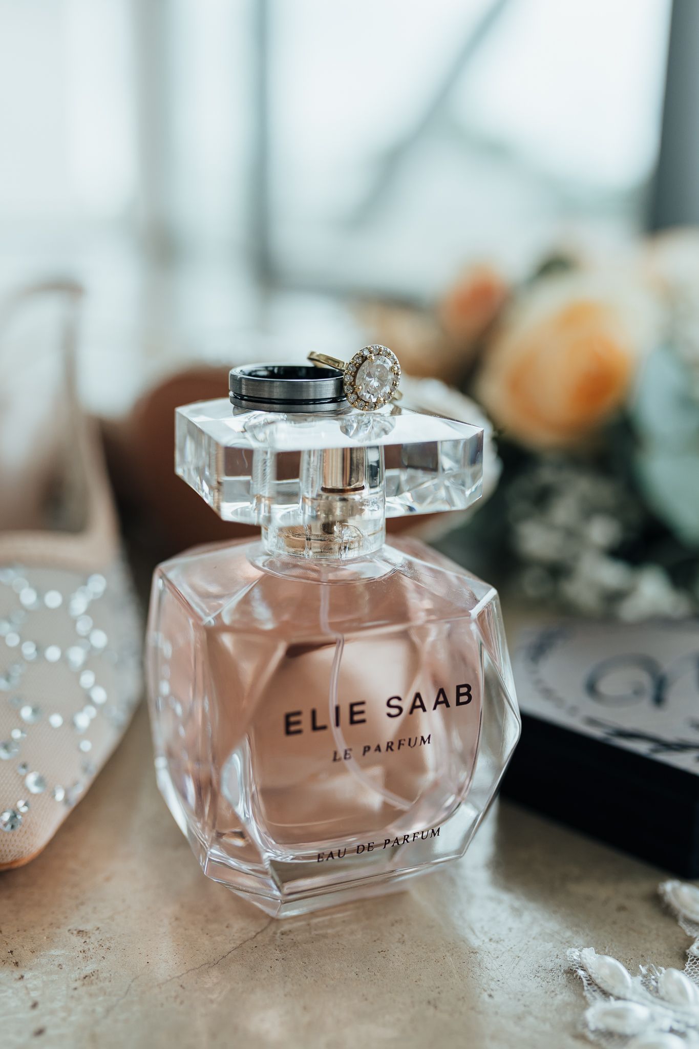 Elie Saab perfume bottle with engagement ring balanced on top near floral decor.