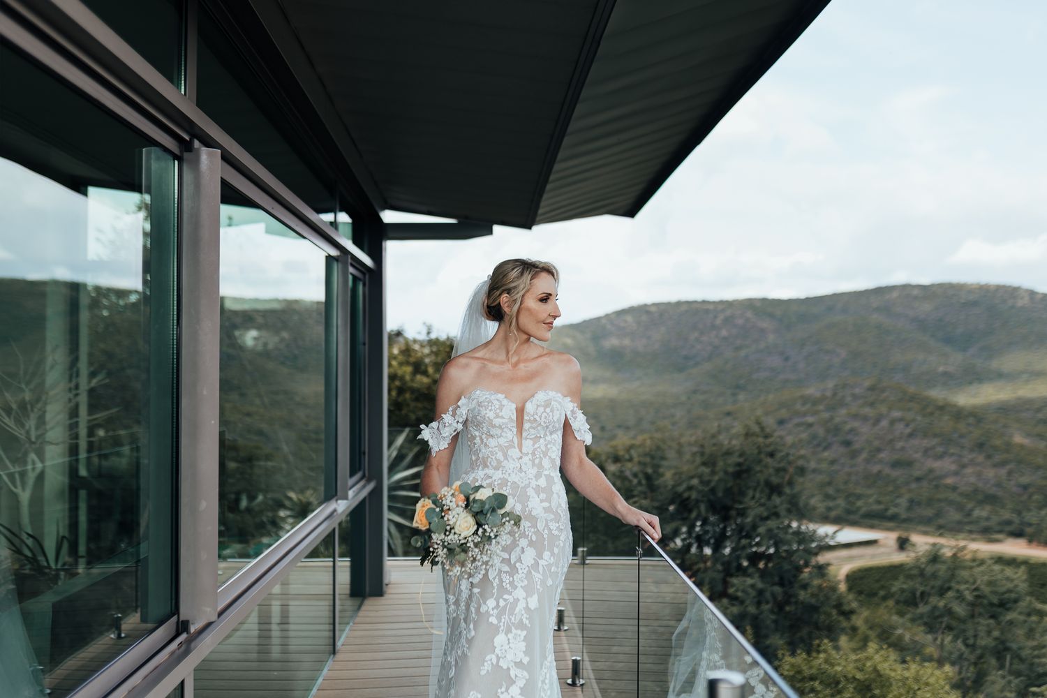 Elegant bride in fitted lace dress poses on contemporary deck with mountain views.
