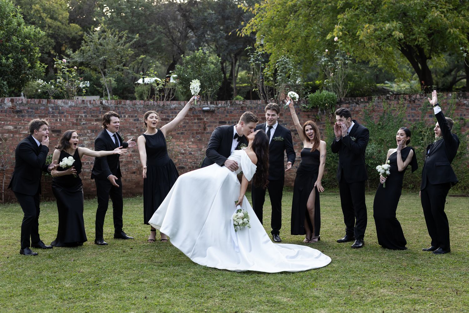 Wedding party members celebrate with fun poses on a lawn while the dress train spreads dramatically.