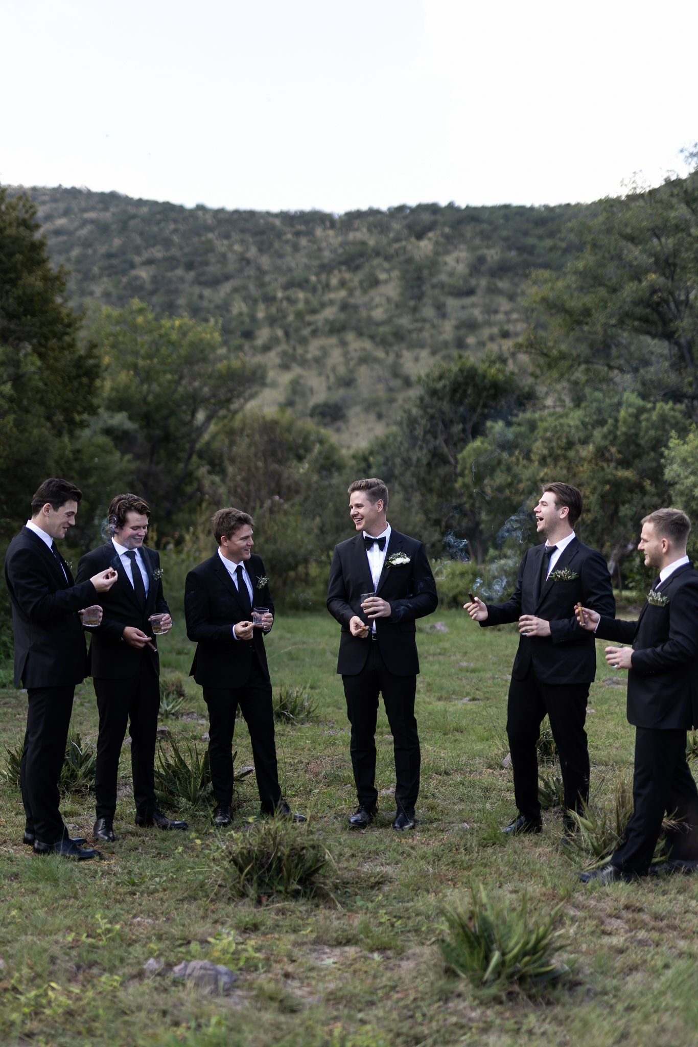 Wedding party in black suits gathered outdoors with rolling hills in the background.