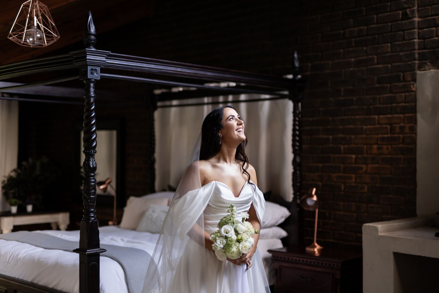 Bride holding white bouquet poses elegantly by four-poster bed with exposed brick walls.