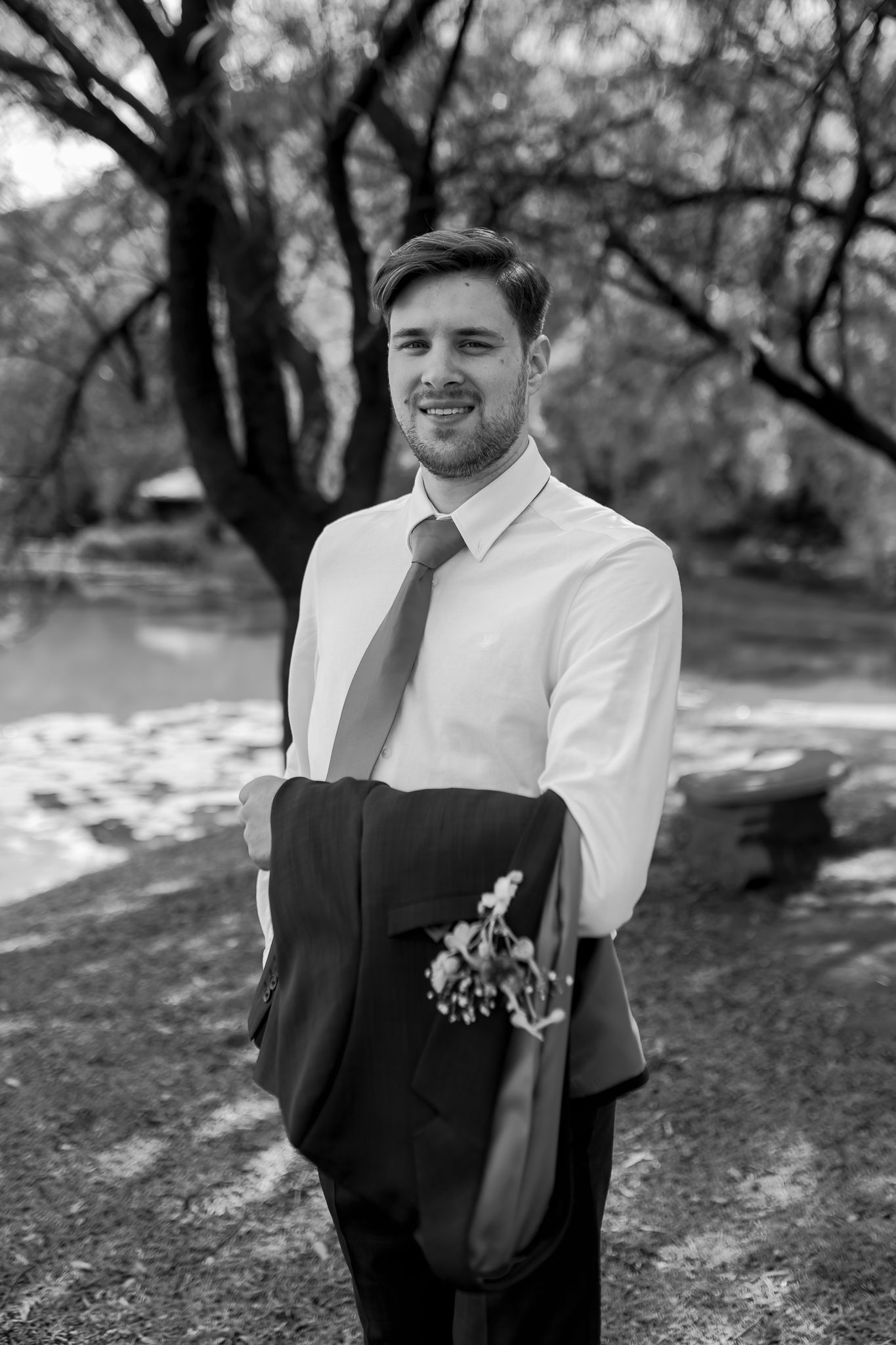 A groom in formal attire holds his suit jacket while posing outdoors in a black and white photo.