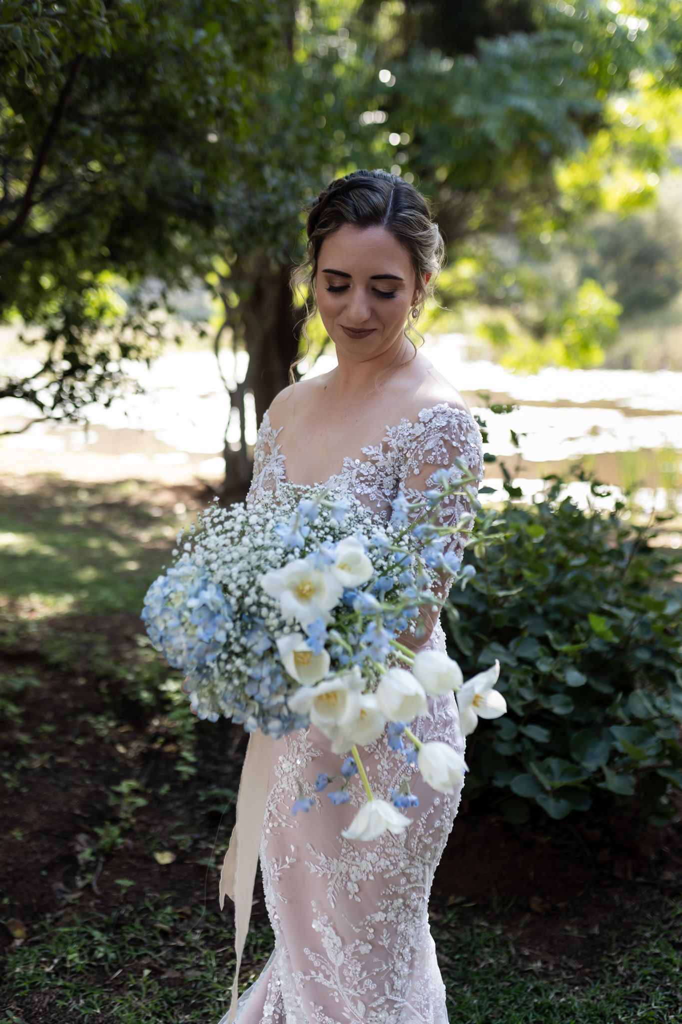 A bride in an embroidered gown holding a bouquet of white calla lilies and baby's breath in a garden.