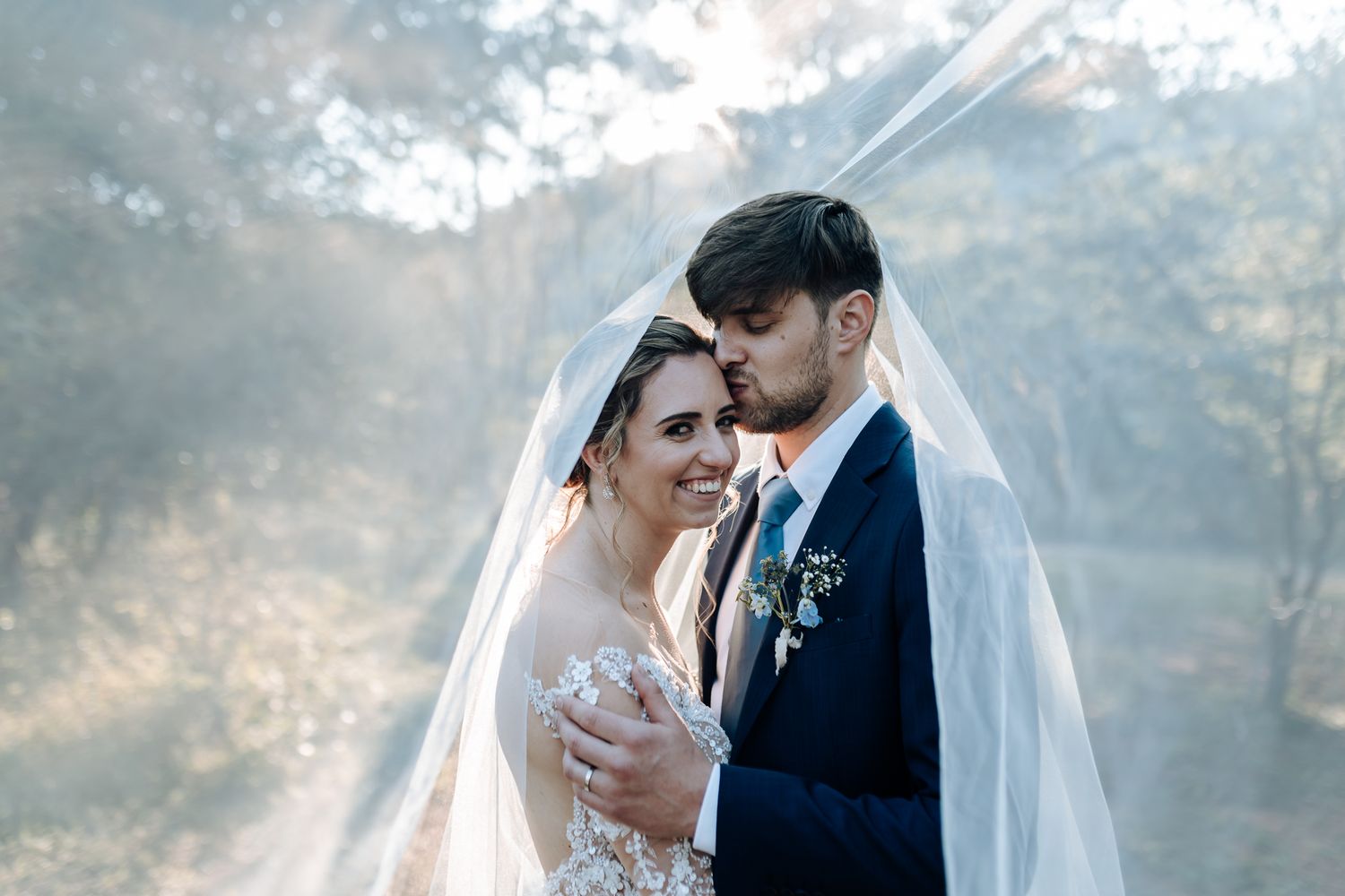 A romantic wedding portrait showing a couple embracing under a dreamy sky with the bride's veil flowing in the wind.