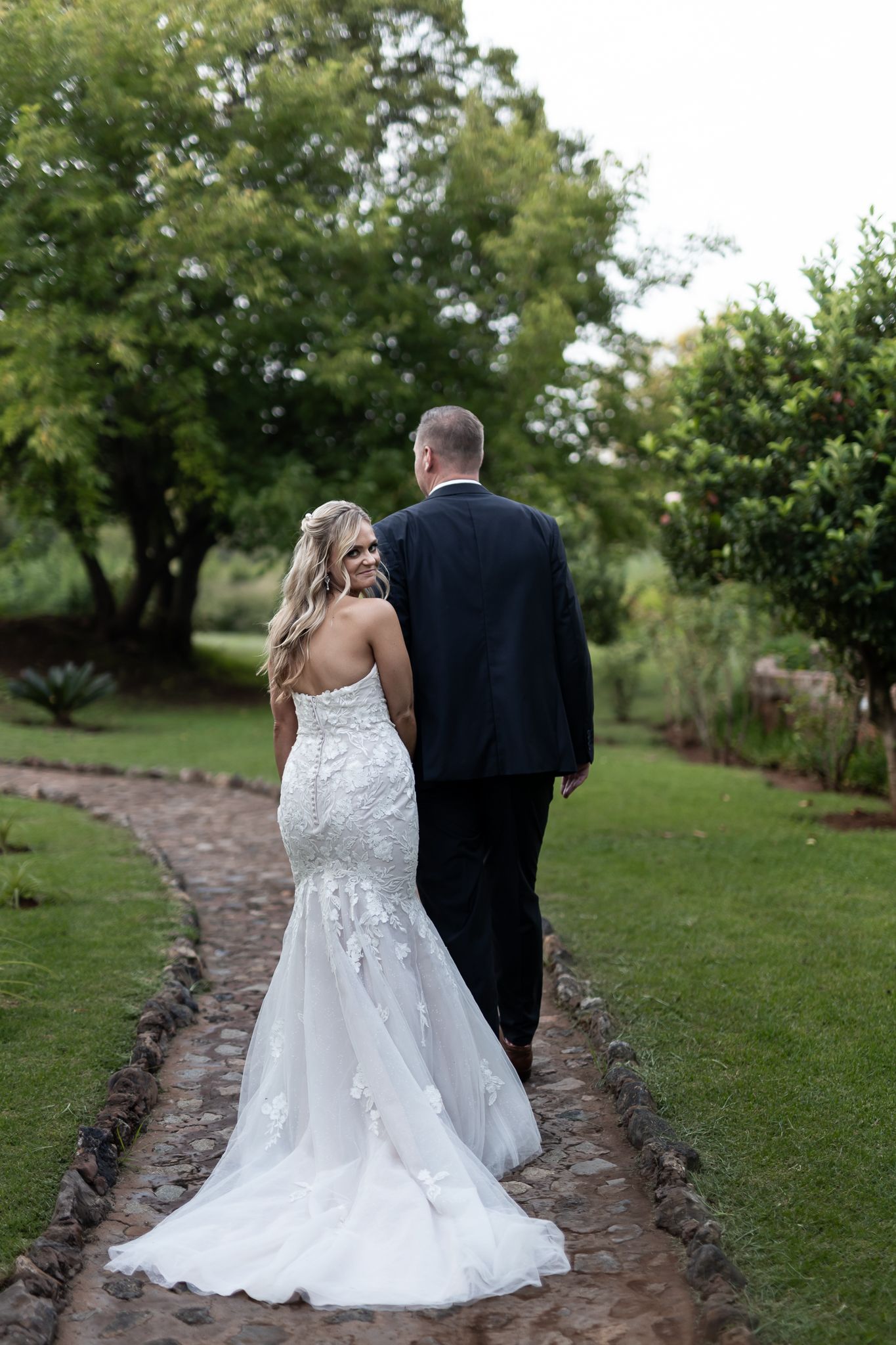 A couple in wedding attire walks along a stone path surrounded by green foliage.